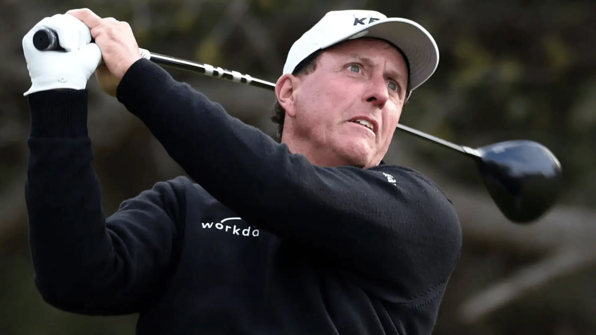 phil mickelson wearing a white cap and black sweater swings a golf club during a shot with a focused expression featured image