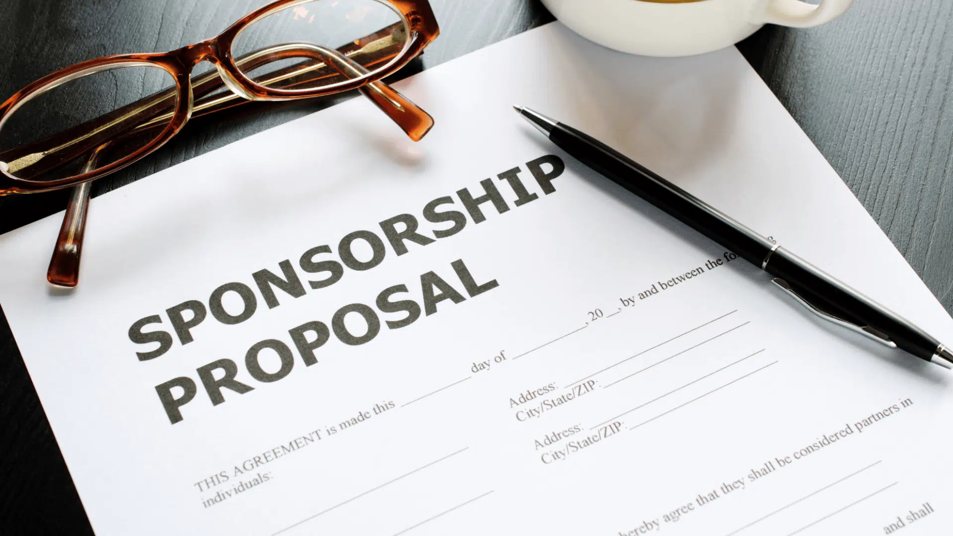 A sponsorship proposal document on a desk with a pen reading glasses and a coffee cup placed beside it ready for review or signing