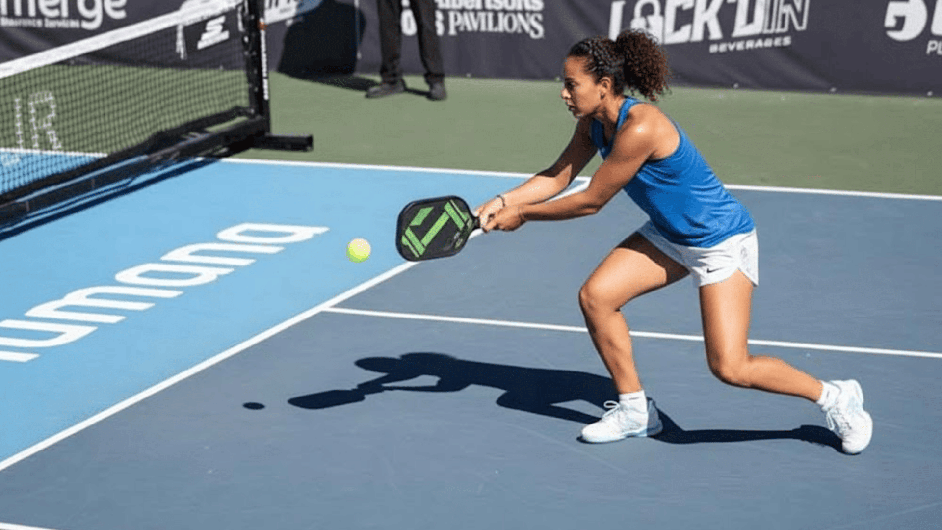 A woman in an athletic wear is playing pickleball lunging towards a ball with a paddle on a sunny court