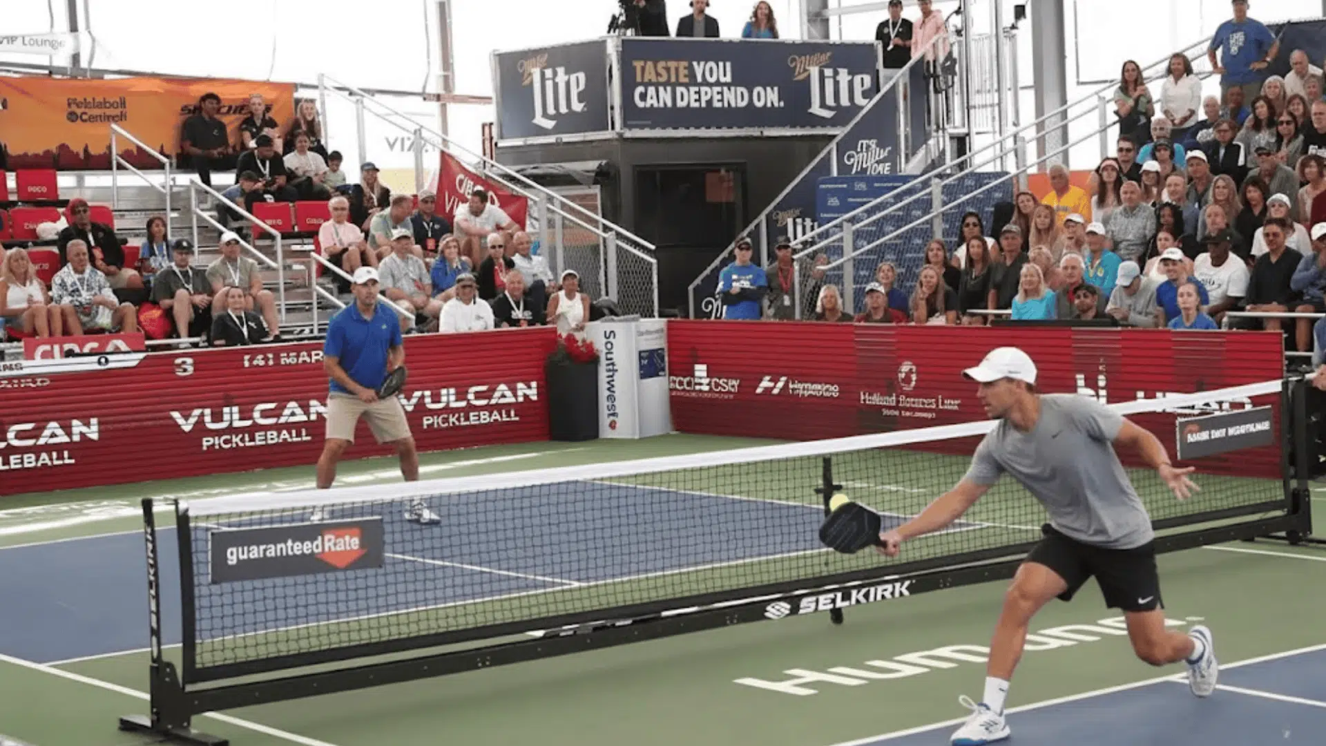 a professional pickleball player reaches forward to hit a shot during a live match with spectators watching from the stands featured image