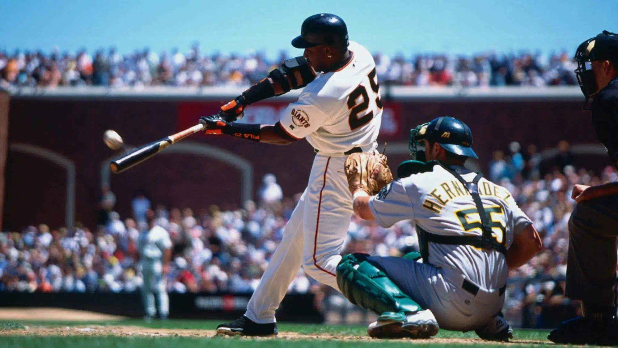 A baseball player swings a bat to hit the ball as a catcher crouches behind home plate with a stadium crowd in the background