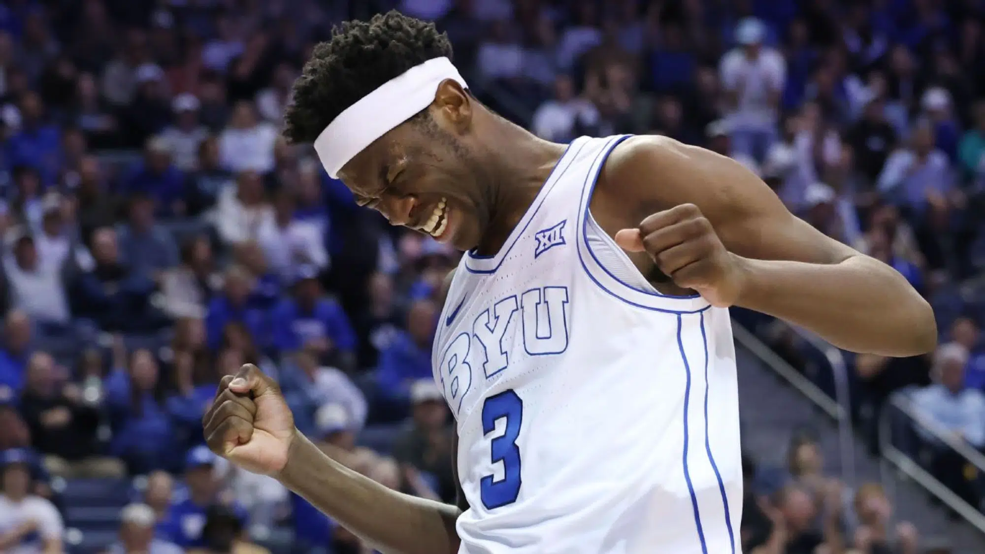 a BYU basketball player wearing number 3 pumps his fists and smiles during a game in a crowded arena
