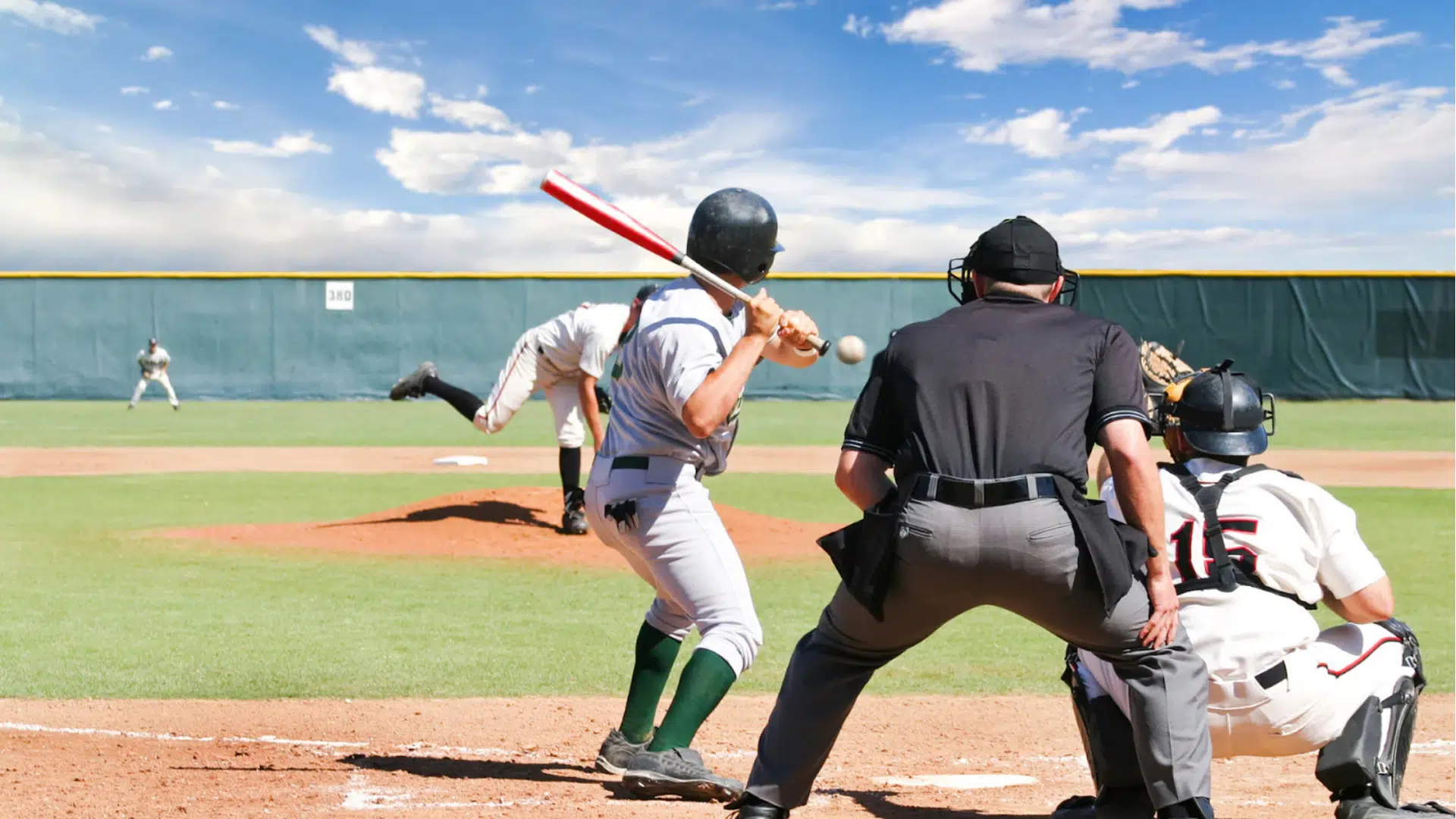a baseball batter preparing to swing at a pitch while the catcher and umpire crouch behind home plate on a sunny field