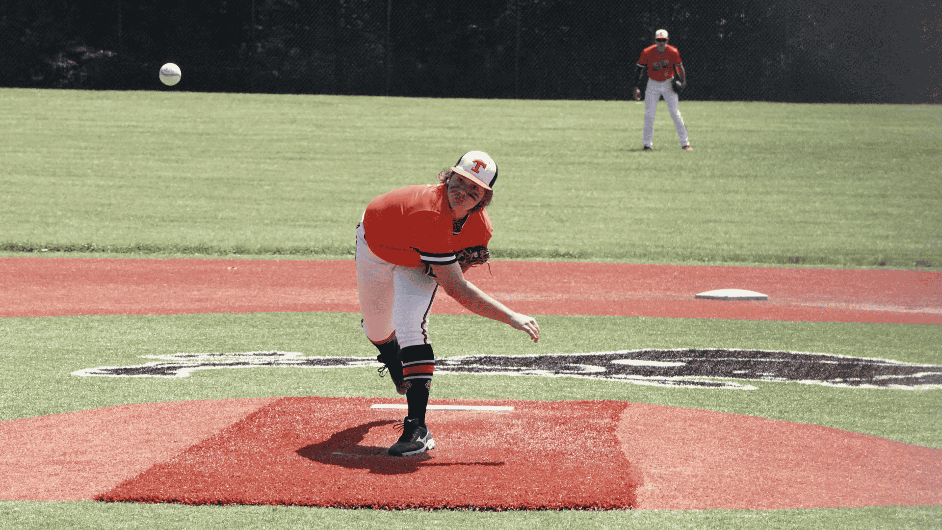 a baseball pitcher in an orange jersey follows through on a pitch on a sunny field