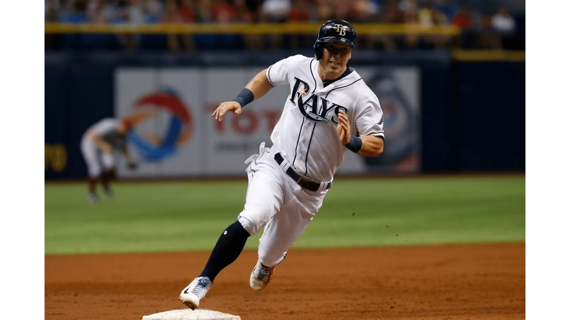 a baseball player in a white rays uniform running past a base during a game on a dirt infield with crowd in background