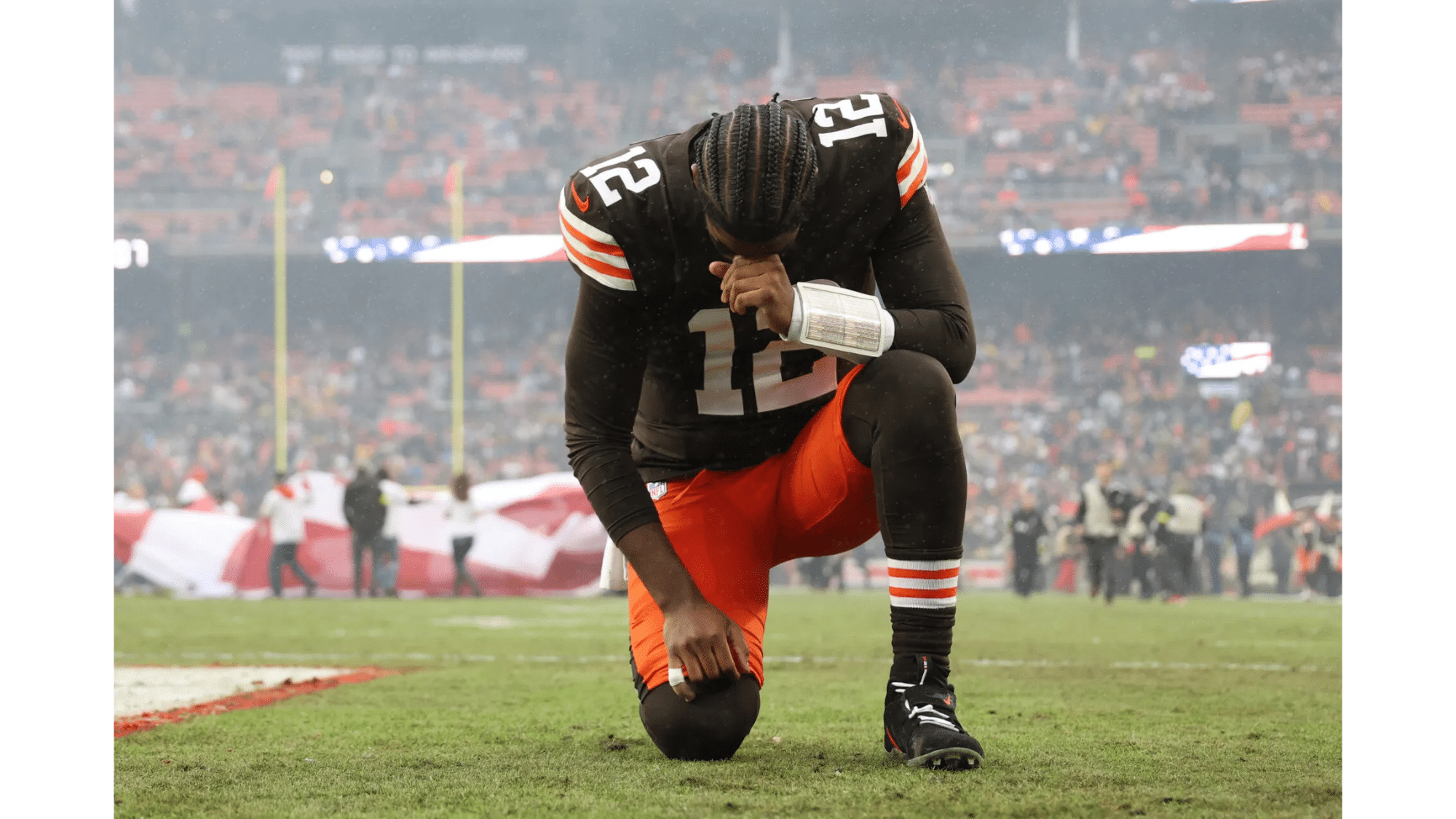 a cleveland browns football player wearing number 12 kneels on the field with his head bowed during a game in a crowded stadium