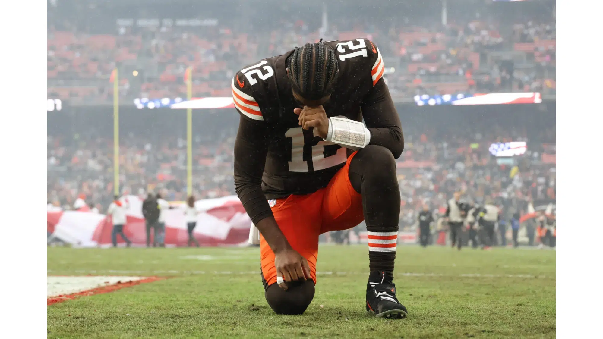 a cleveland browns football player wearing number 12 kneels on the field with his head bowed during a game in a crowded stadium