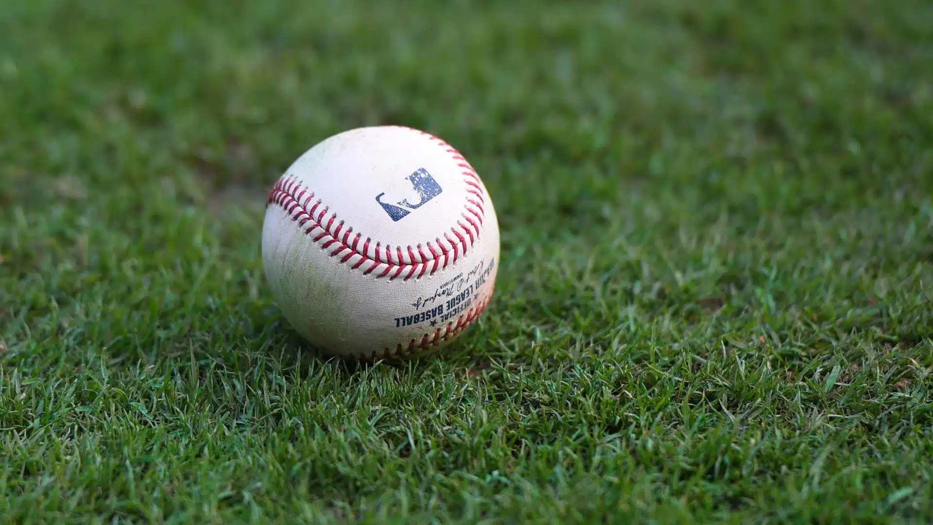 a close up of a worn baseball resting on green grass showing red stitching and scuff marks from repeated use in play