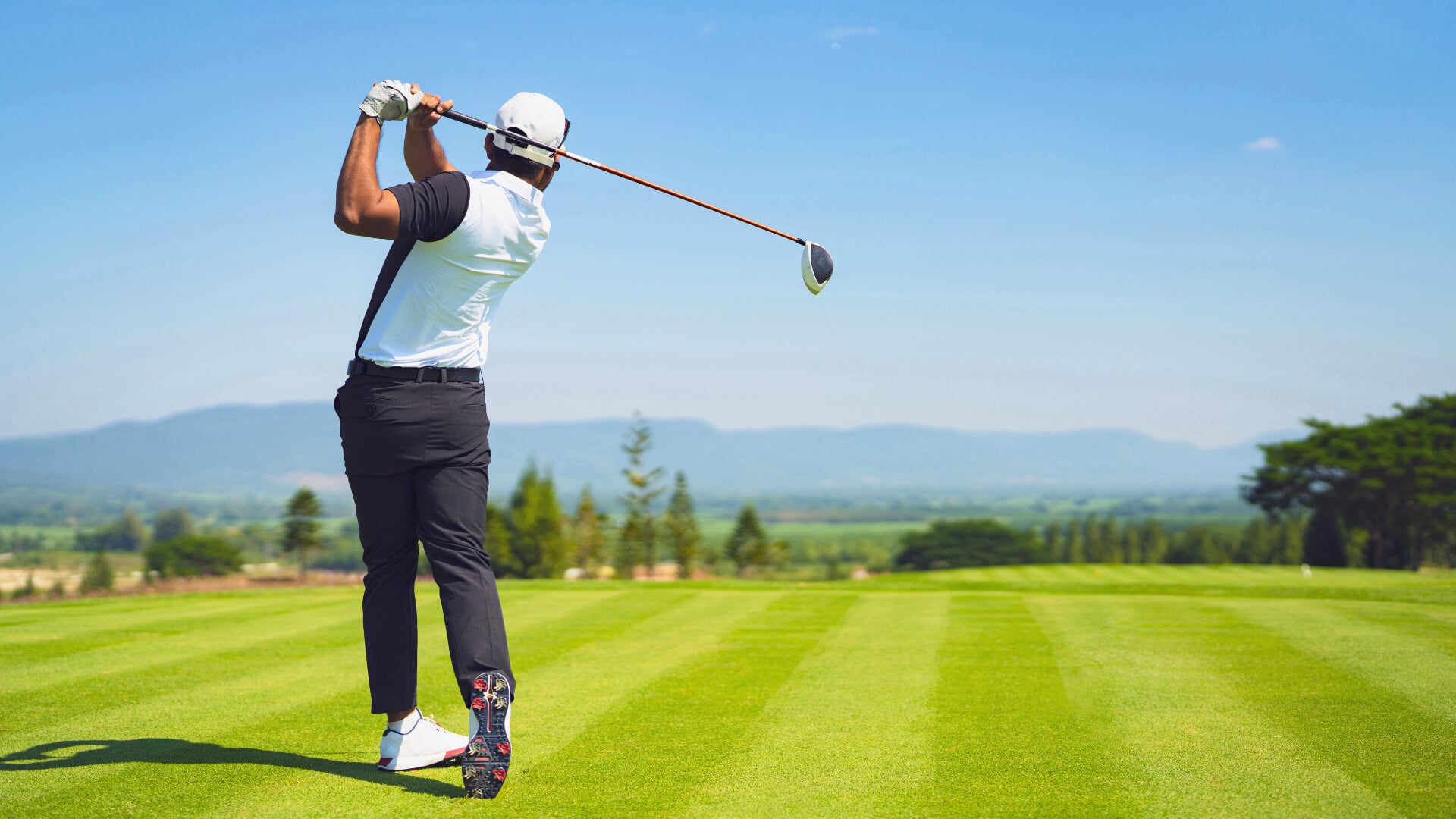 a golfer completes a full driver follow through on a wide open course with rolling hills and mountains in the distance