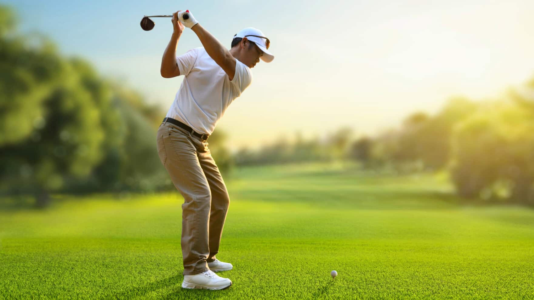 a golfer stands on a lush green fairway at sunrise completing a backswing with a driver while focusing on a teed golf ball
