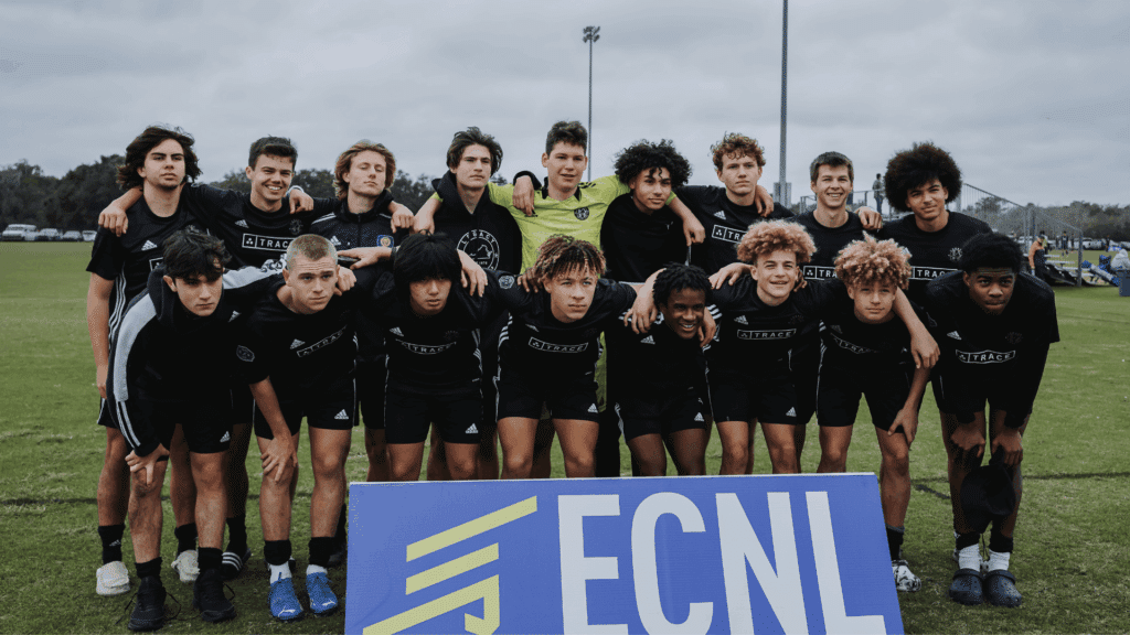 a group of teenage boys in black soccer uniforms posing together on a grassy field behind an ECNL sign under a cloudy sky