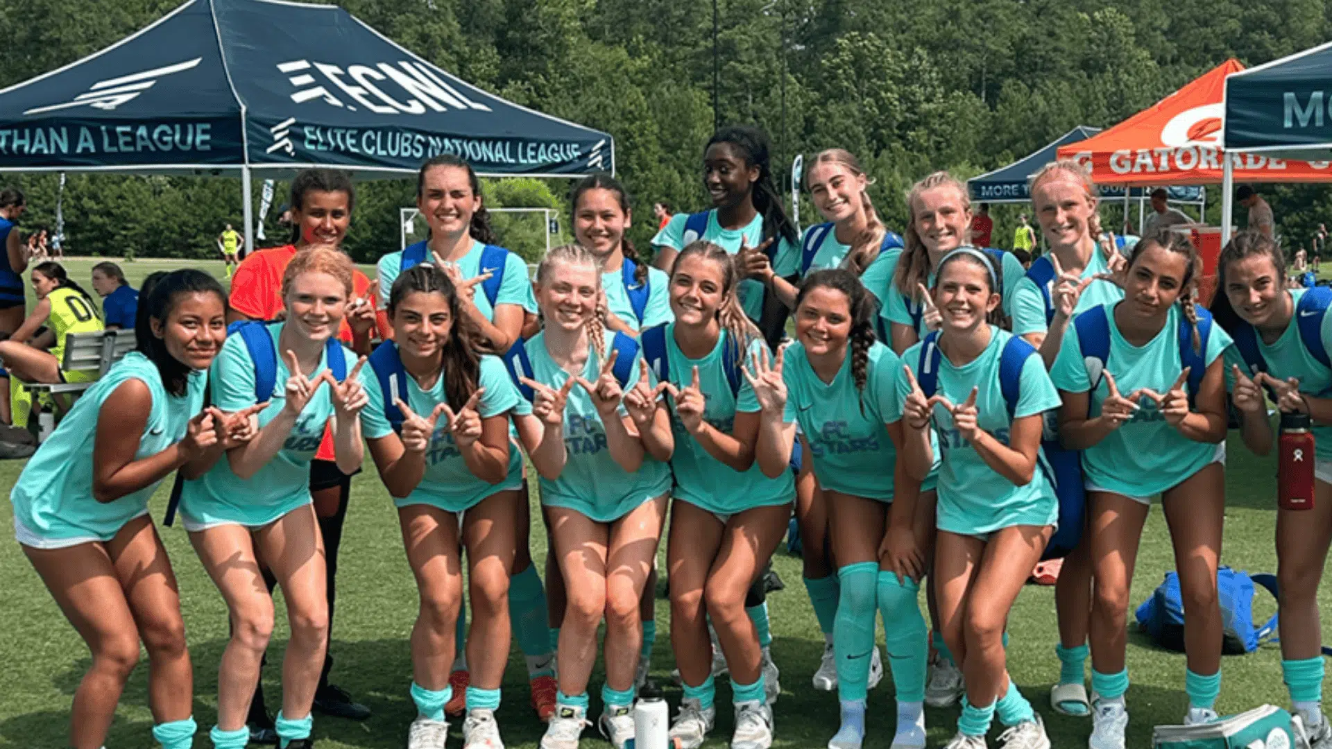 a group of teenage girls in teal soccer uniforms posing together on a field at ECNL Finals with event tents in the background