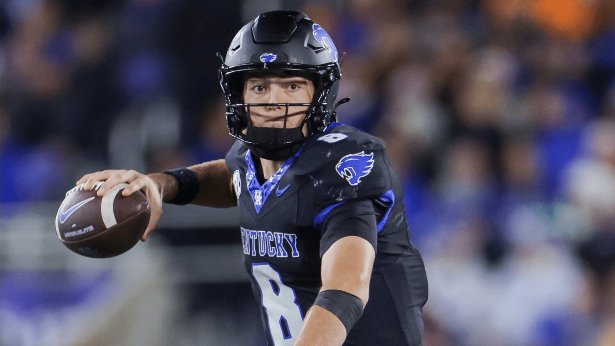 a kentucky football player wearing number 8 prepares to throw a pass during a night game with a blurred crowd in the background