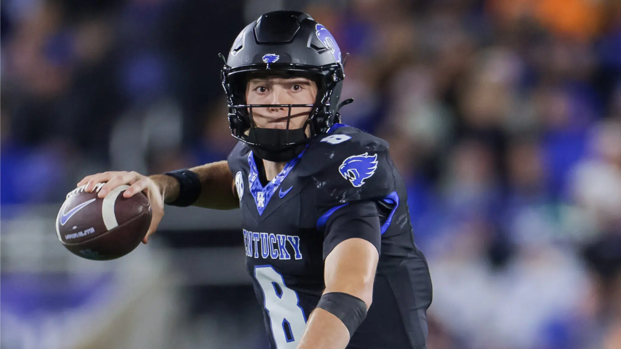 a kentucky football player wearing number 8 prepares to throw a pass during a night game with a blurred crowd in the background