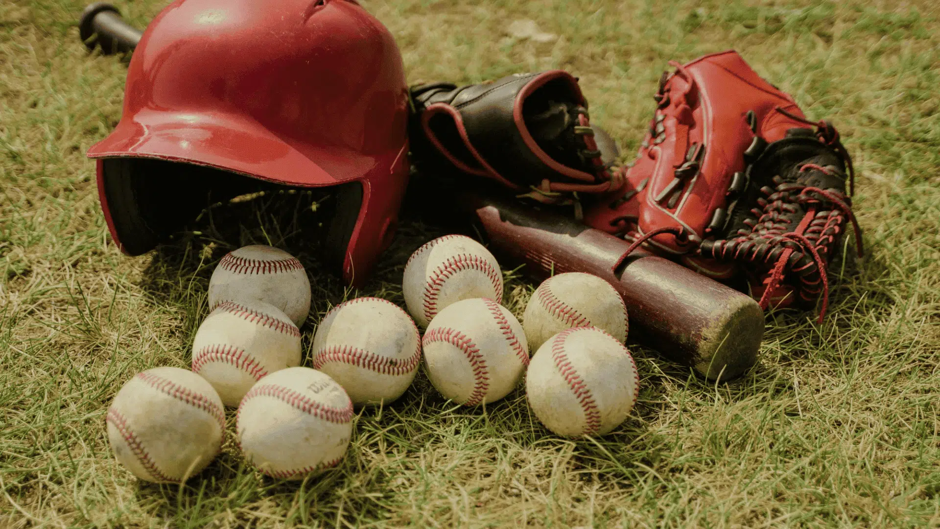 a red baseball helmet bat glove and several baseballs resting together on the grass