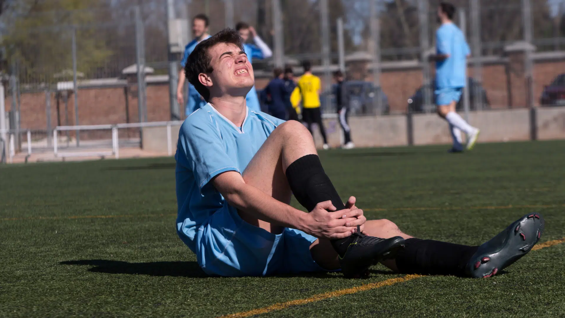 a soccer player in a blue uniform sits on the field clutching his knee in pain during a match while teammates play behind him