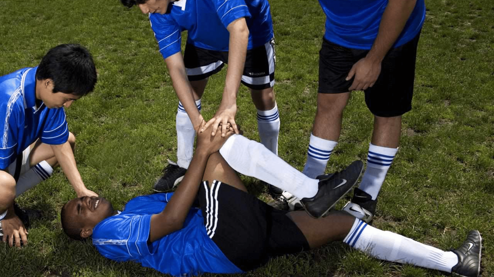 a soccer player lies on the grass holding his leg in pain while teammates in blue uniforms assist him during a match