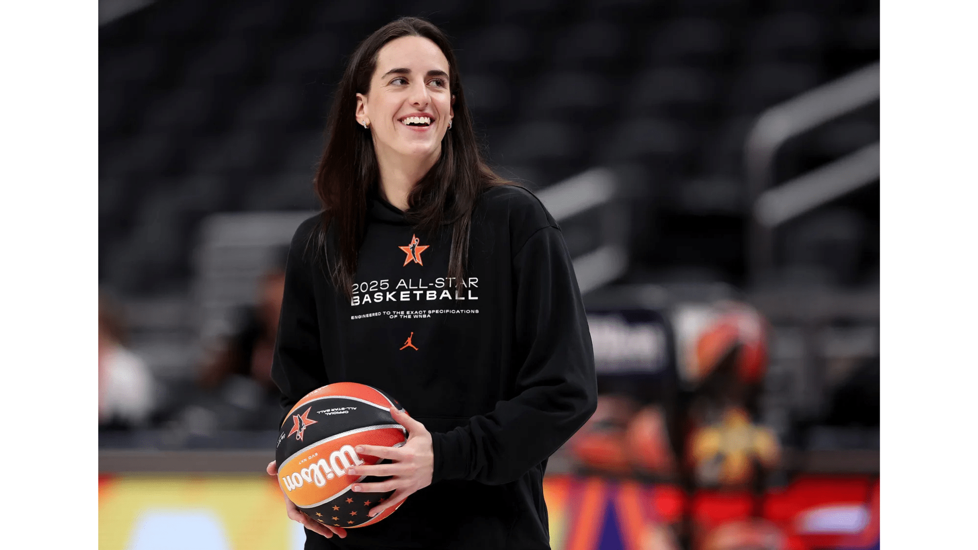 a top nil earners wearing a black 2025 all star basketball hoodie smiles while holding a basketball on an indoor court