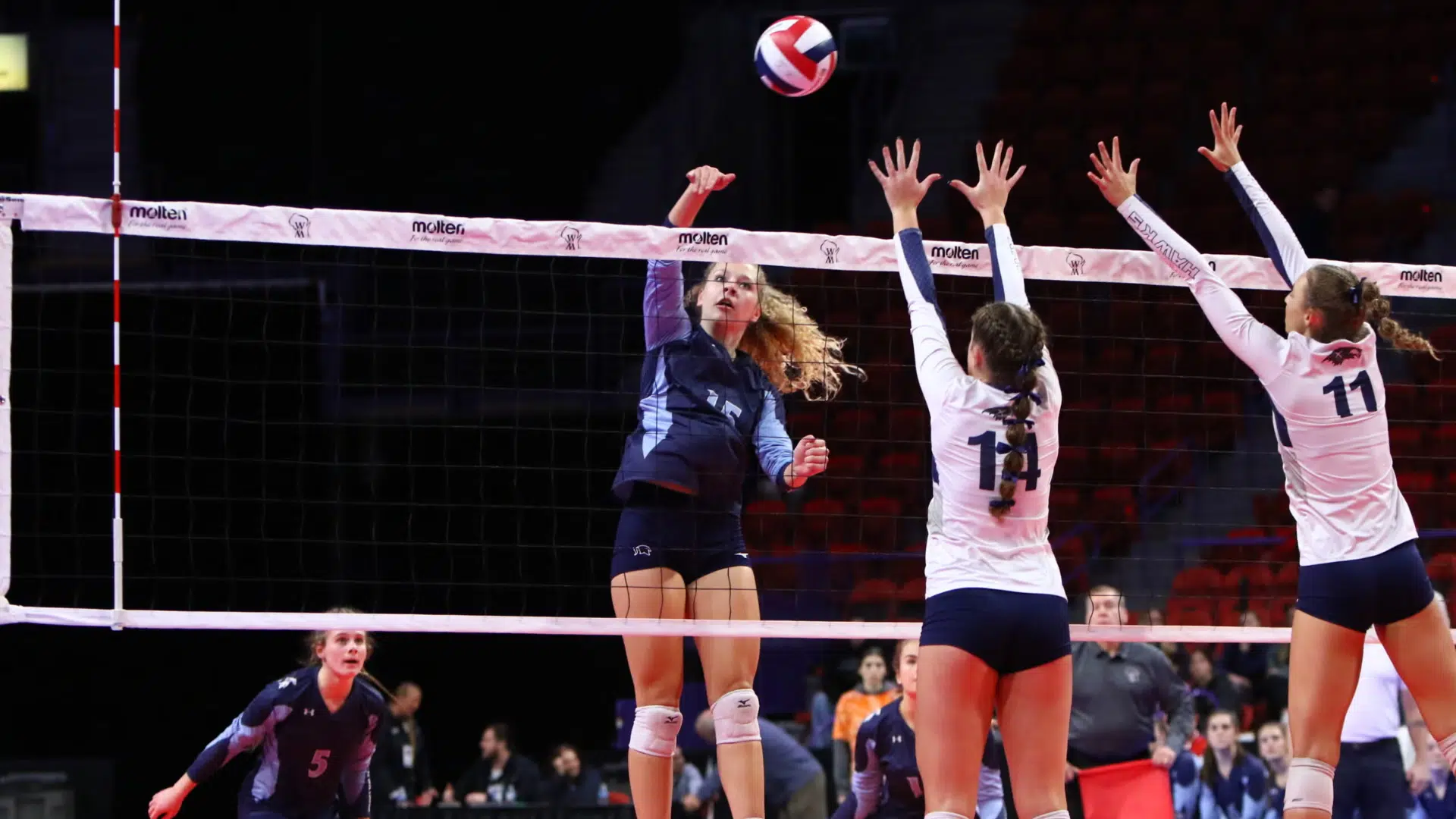 a volleyball player spiking the ball over the net during an indoor match as two blockers jump to defend at the net
