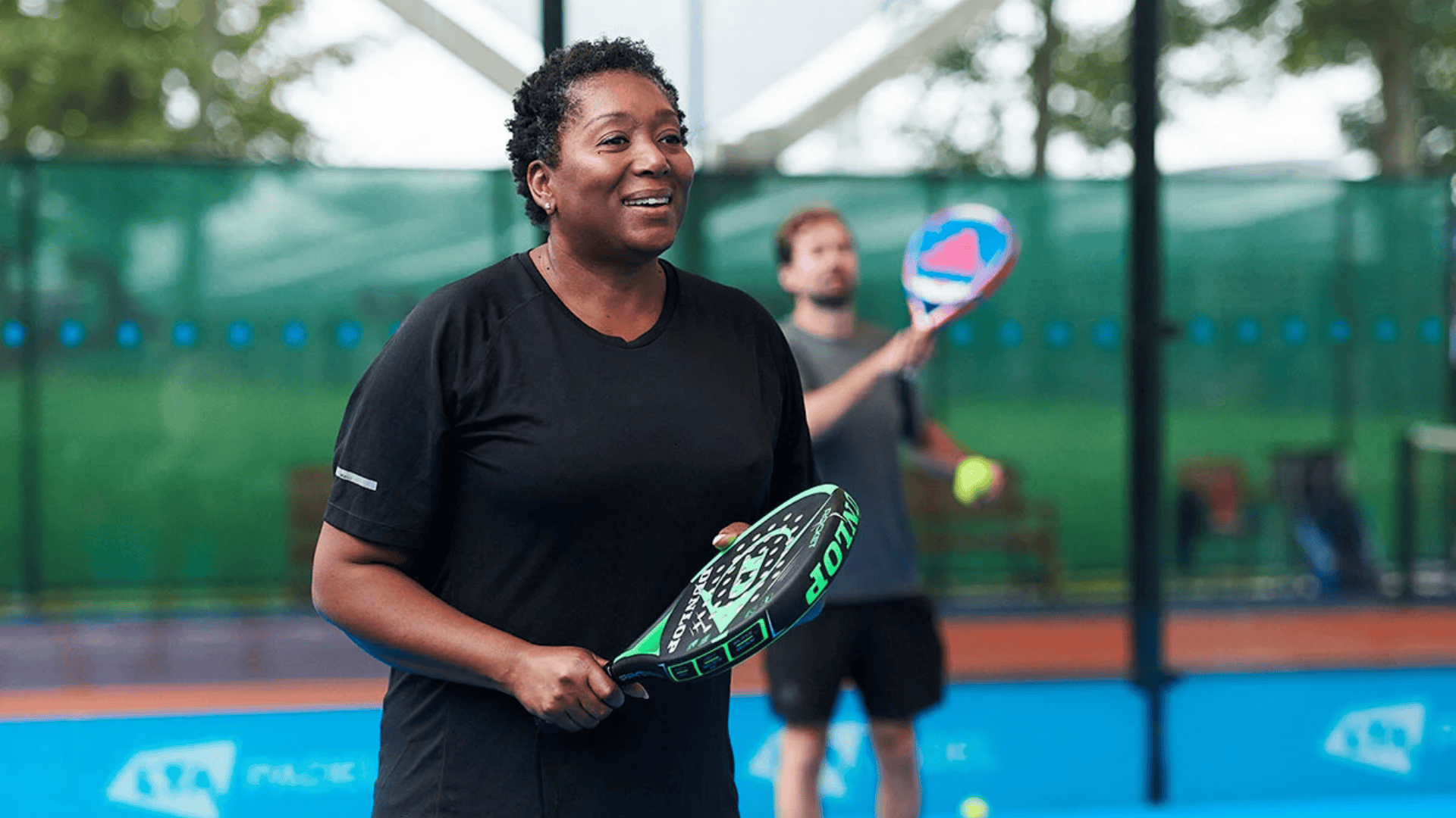 a woman holding a padel racket on an outdoor court while a man prepares to hit a ball in the background