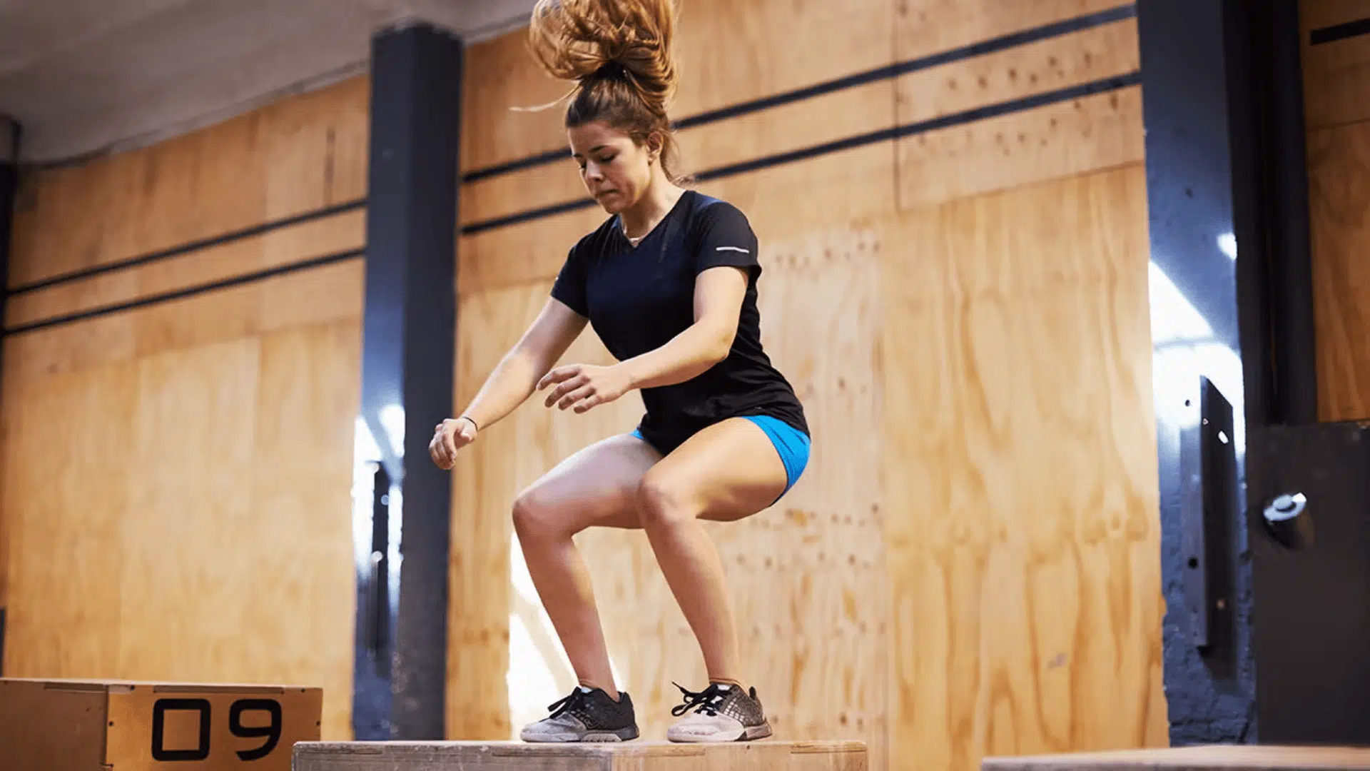 a woman performing a box jump exercise in a gym wearing a black shirt and blue shorts with wooden walls behind her