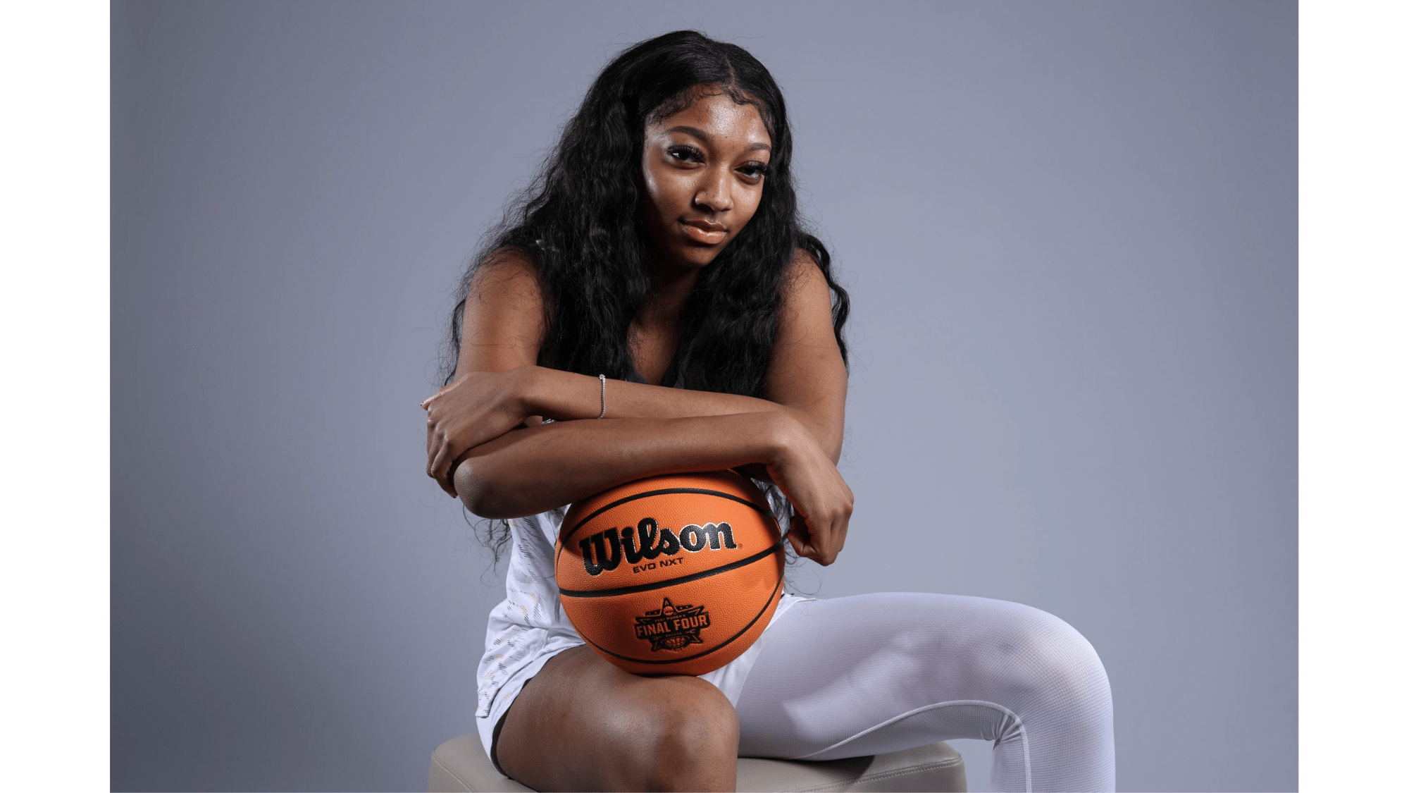 a woman sitting on a stool holding a basketball and looking at the camera against a plain gray background