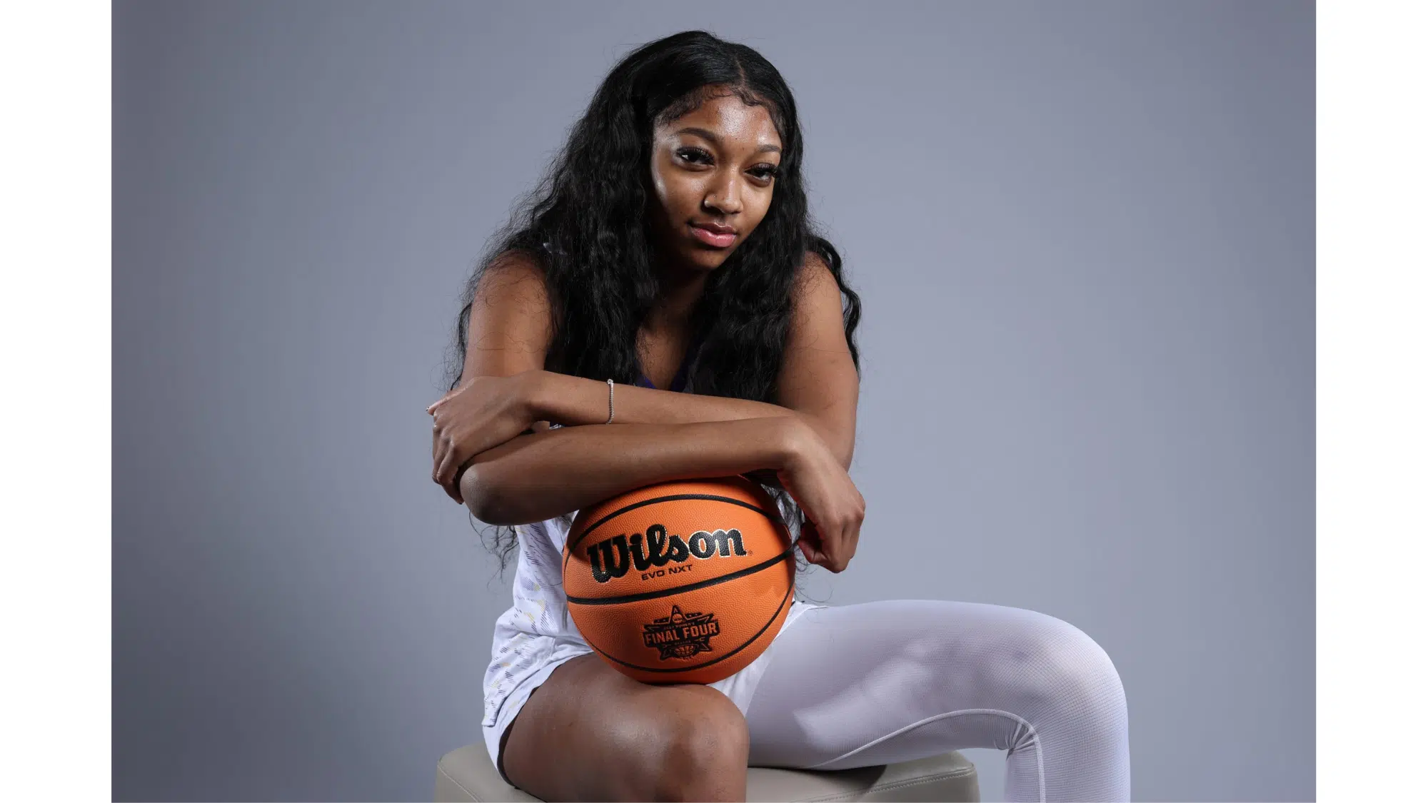 a woman sitting on a stool holding a basketball and looking at the camera against a plain gray background