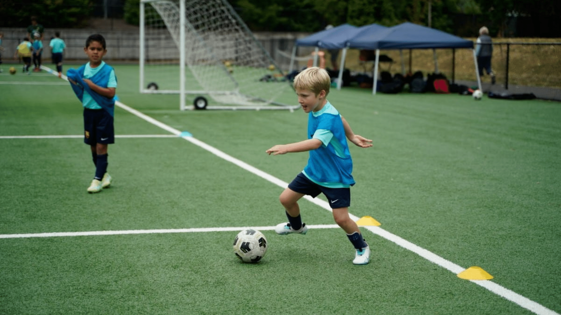 a young boy kicking a soccer ball on a turf field during practice with cones and teammates in the background