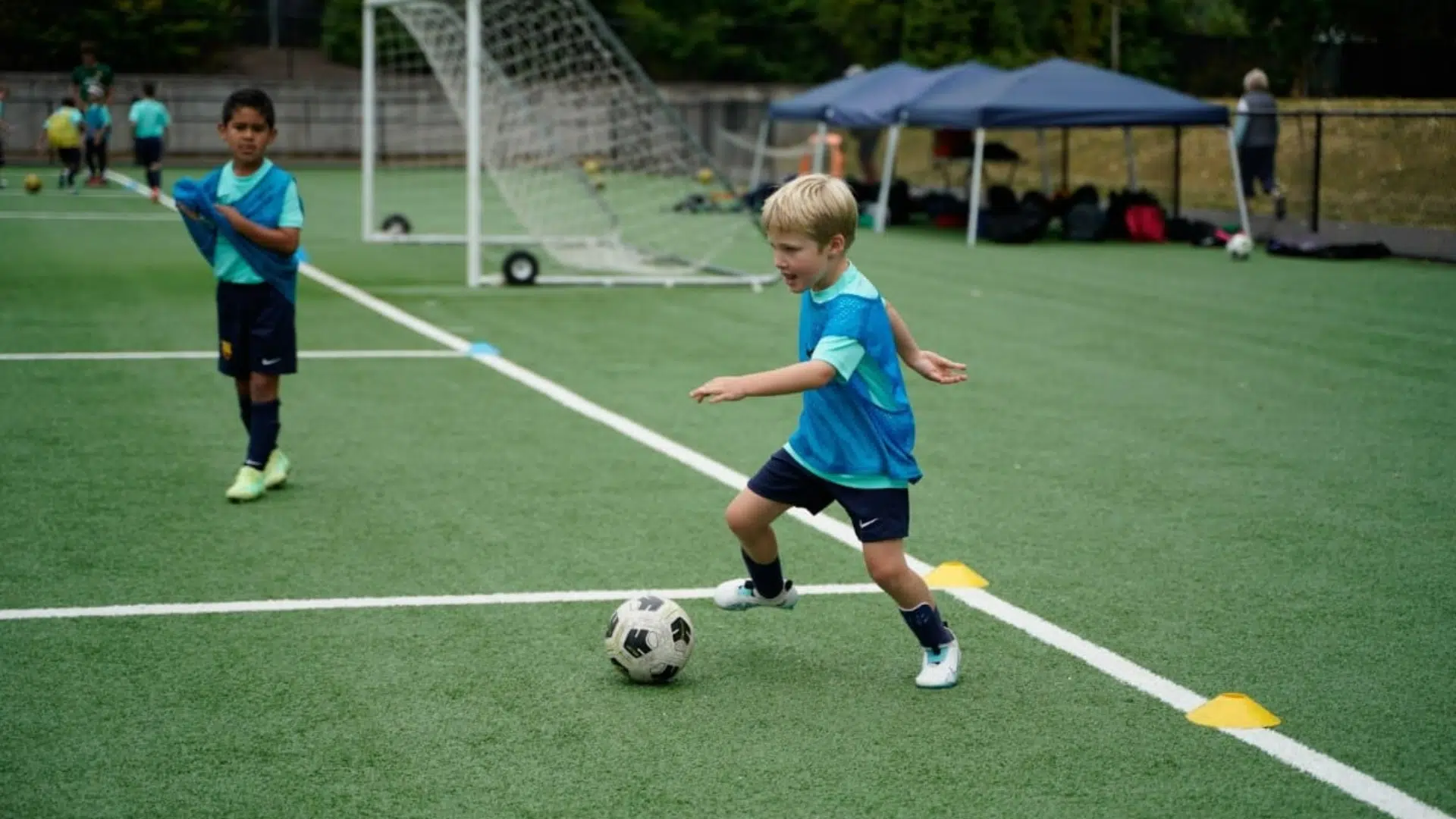 a young boy kicking a soccer ball on a turf field during practice with cones and teammates in the background