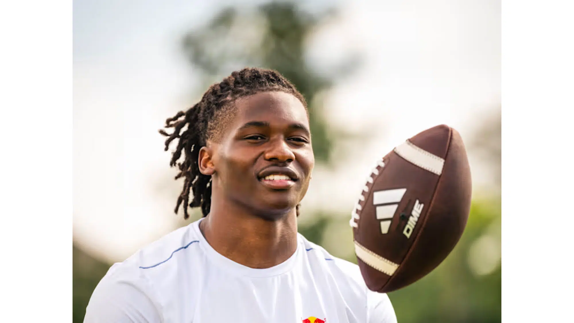 a young top nil earners with dreadlocks smiles as a football spins in the air beside him outdoors on a sunny day