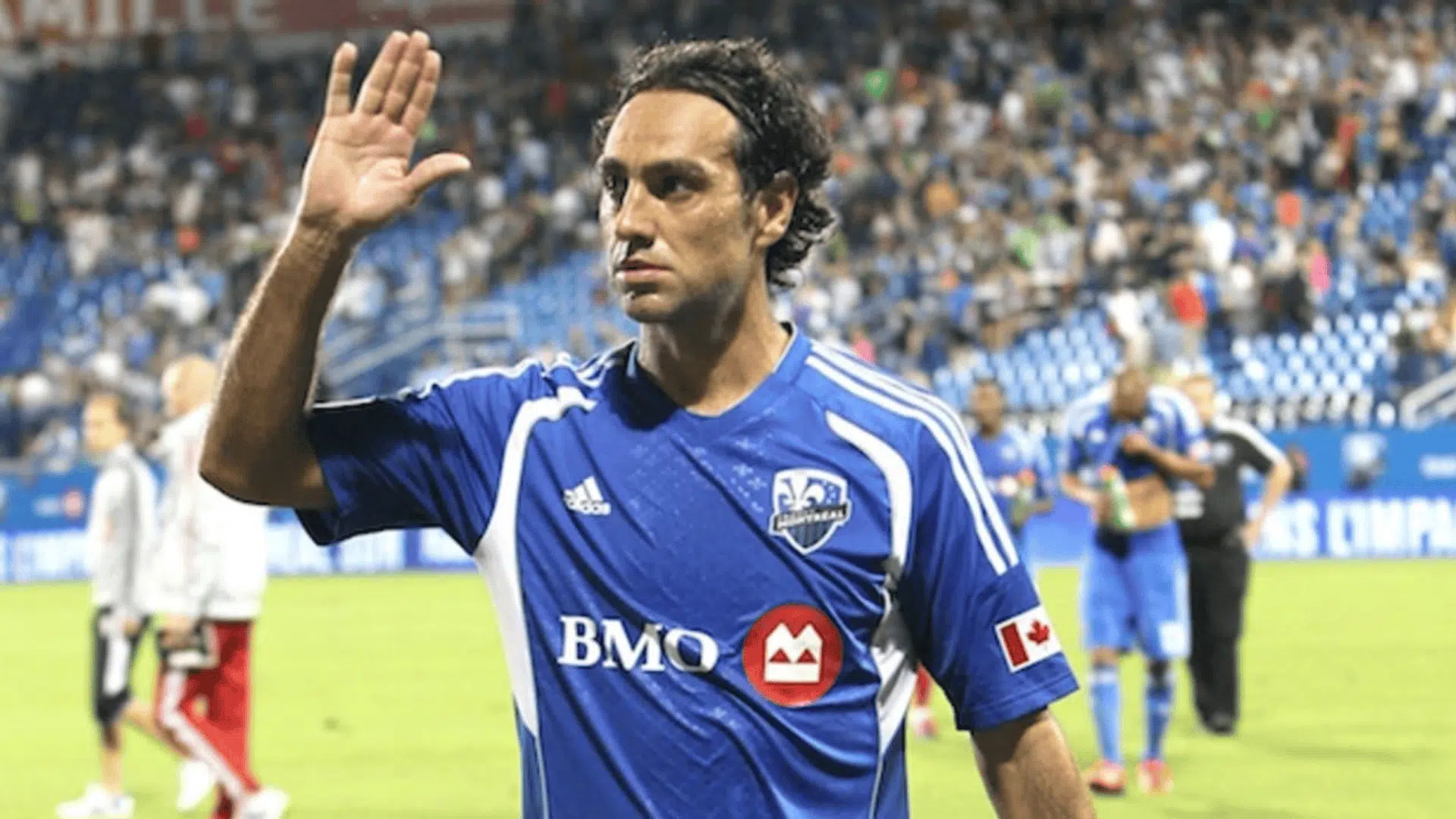 alessandro nesta raising his hand while wearing a blue montreal Impact jersey on a soccer field with a crowd in the background