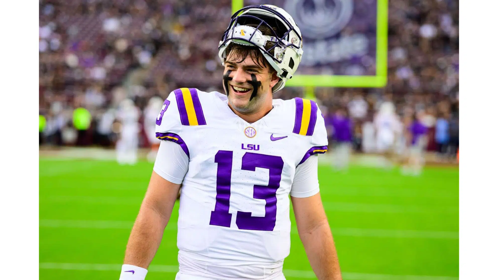 an lsu football player top nil earner wearing number 13 smiles on the field with his helmet raised during a game in a crowded stadium