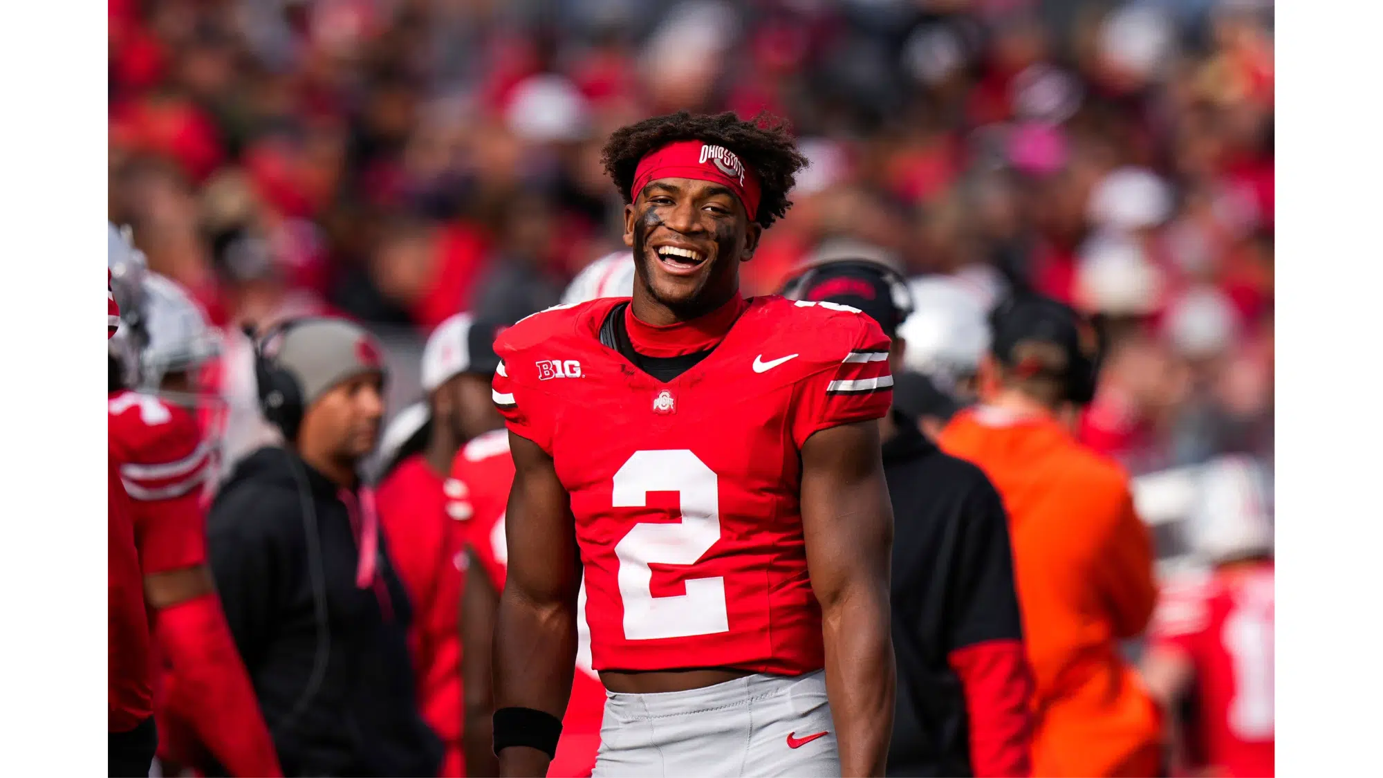 an ohio state football top nil earner player wearing number 2 smiles on the field during a game with teammates and fans in the background