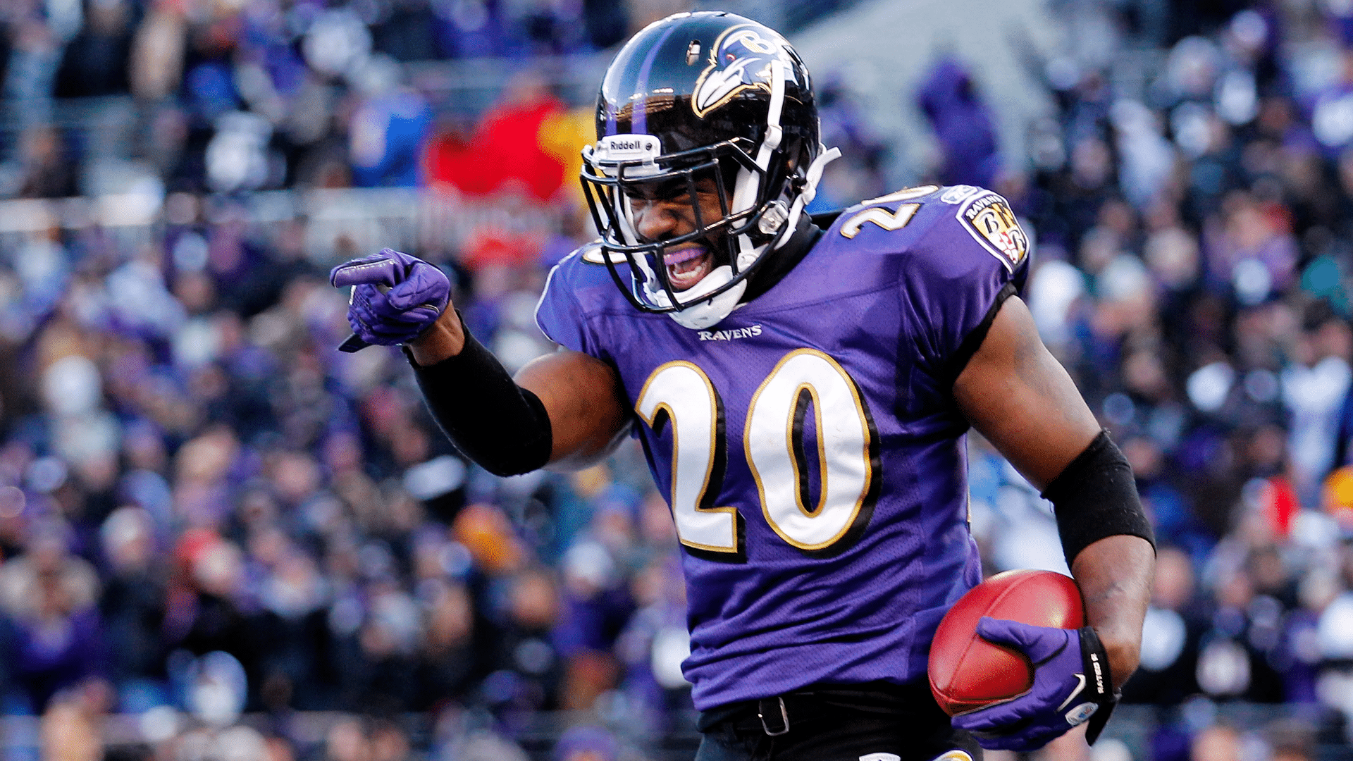 ed reed in a purple uniform holding the ball and pointing while celebrating on the field with a blurred crowd behind him