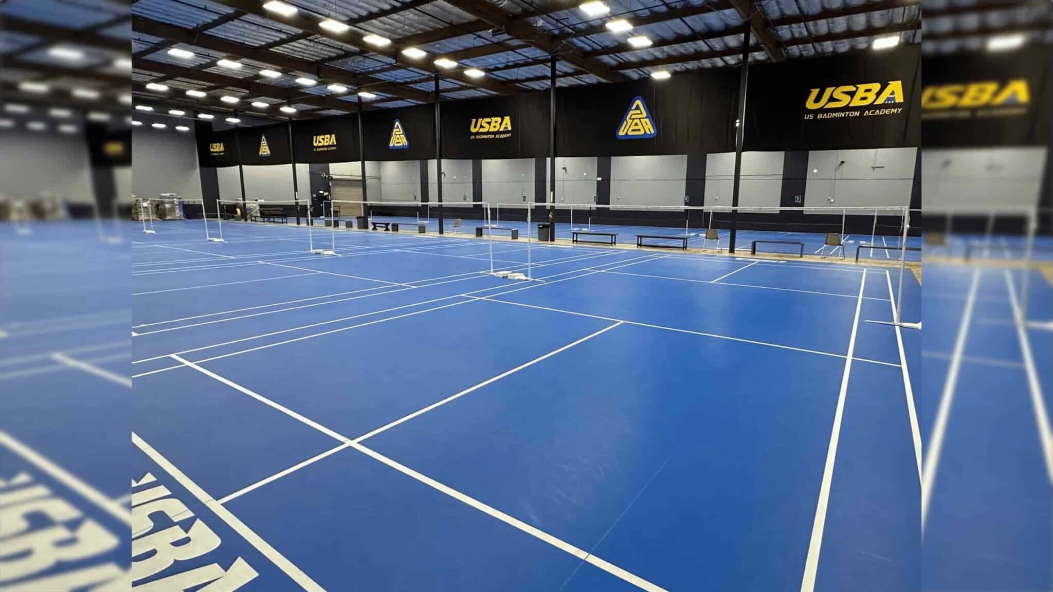 empty indoor badminton courts with blue flooring and usba logos displayed on black walls under bright ceiling lights