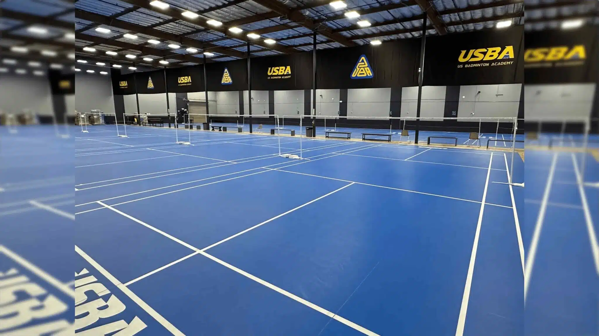 empty indoor badminton courts with blue flooring and usba logos displayed on black walls under bright ceiling lights