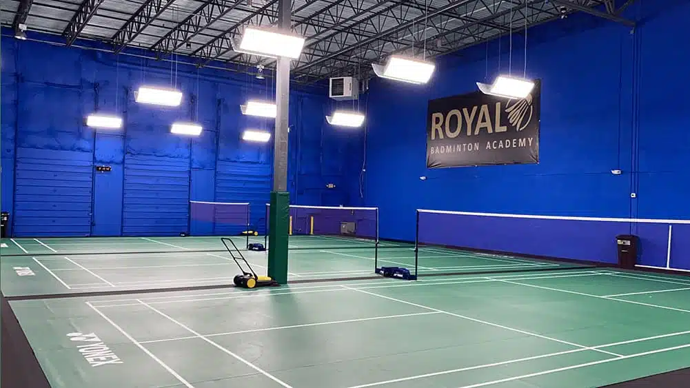 empty indoor badminton courts with green flooring blue walls bright overhead lights and a royal badminton academy sign on the wall