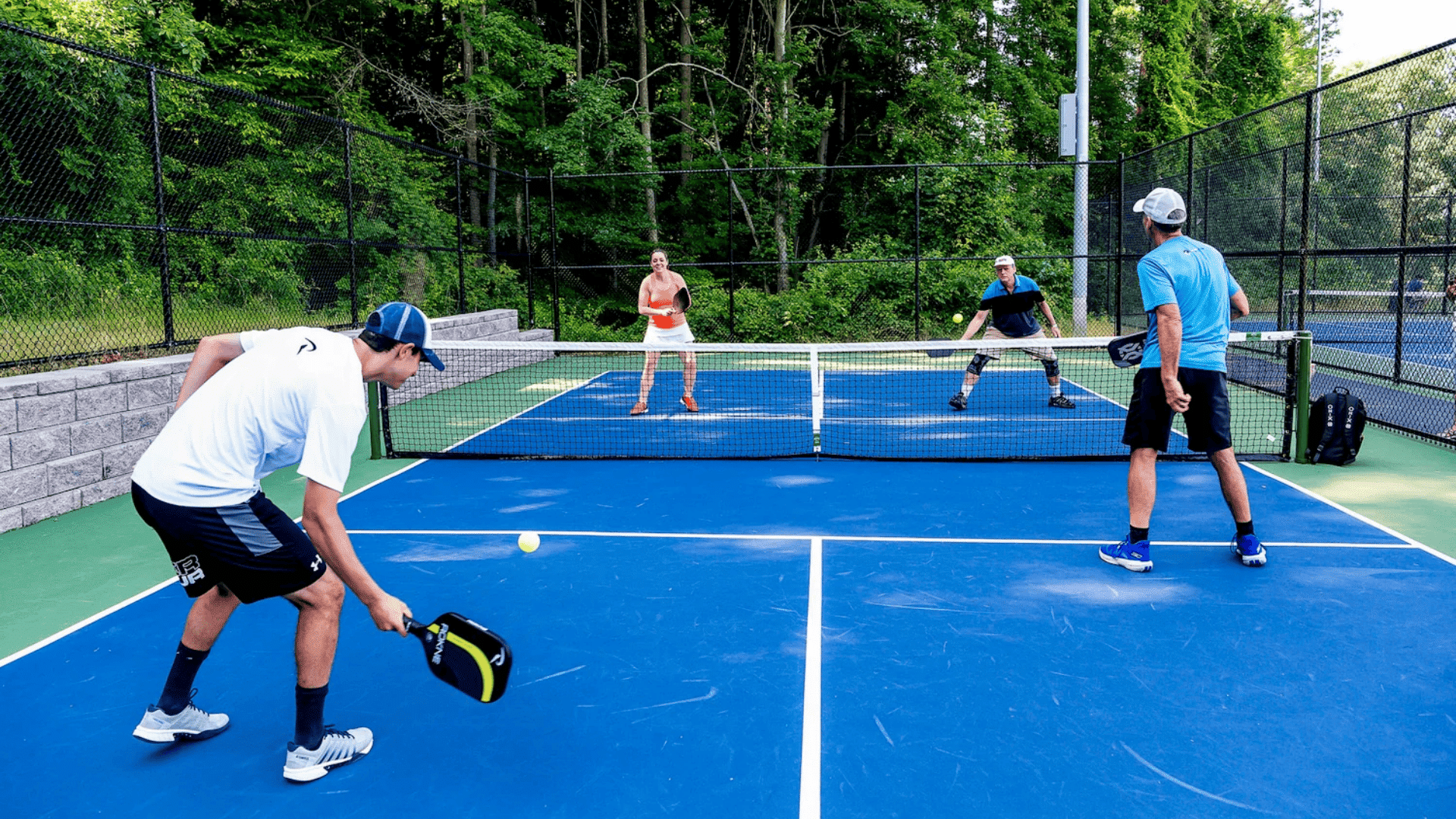four adults playing pickleball on an outdoor court surrounded by trees during a doubles match (1)