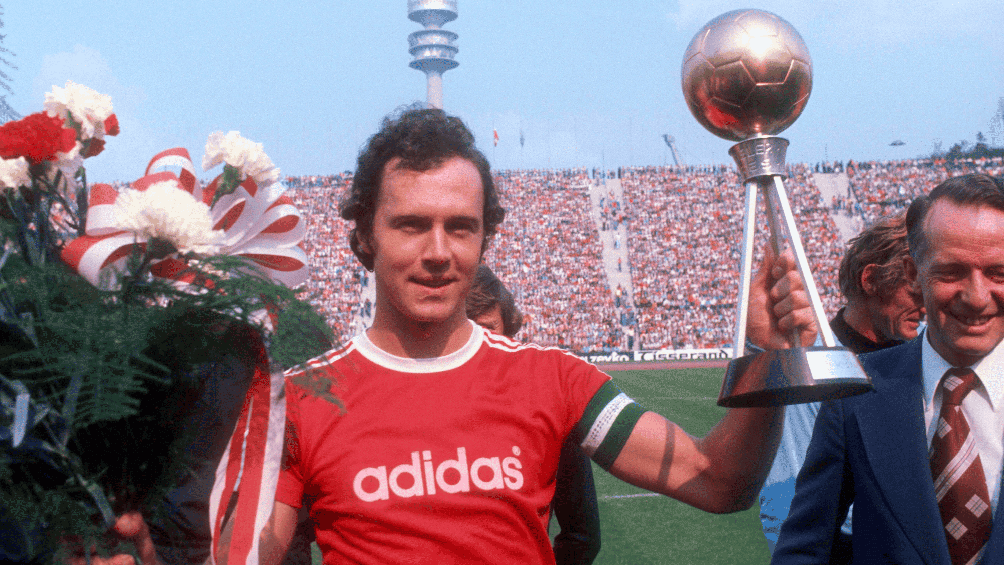 franz beckenbauer holding a trophy and bouquet of flowers in a crowded stadium