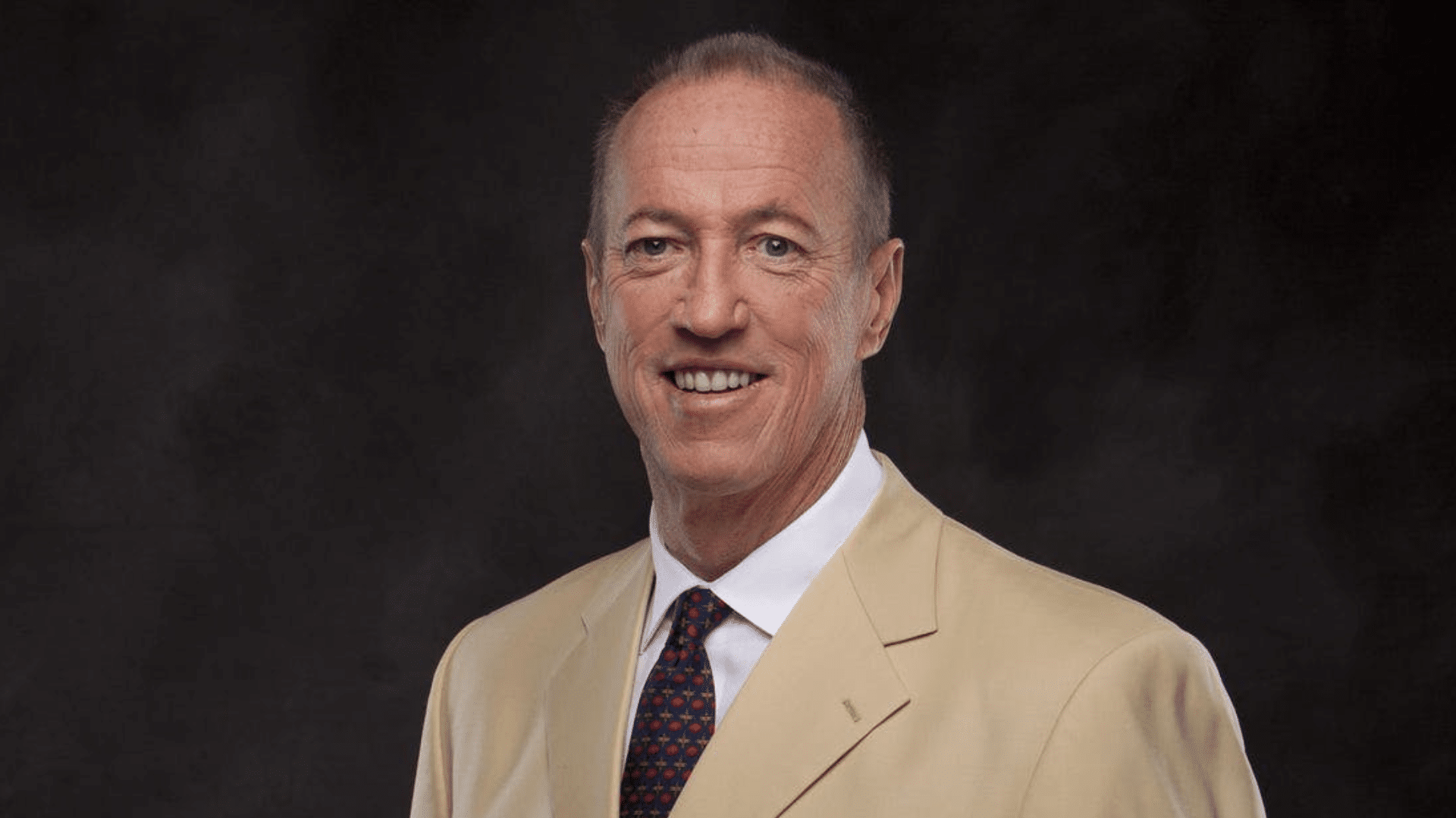 jim kelly smiling man in a light coloured suit poses against a dark background