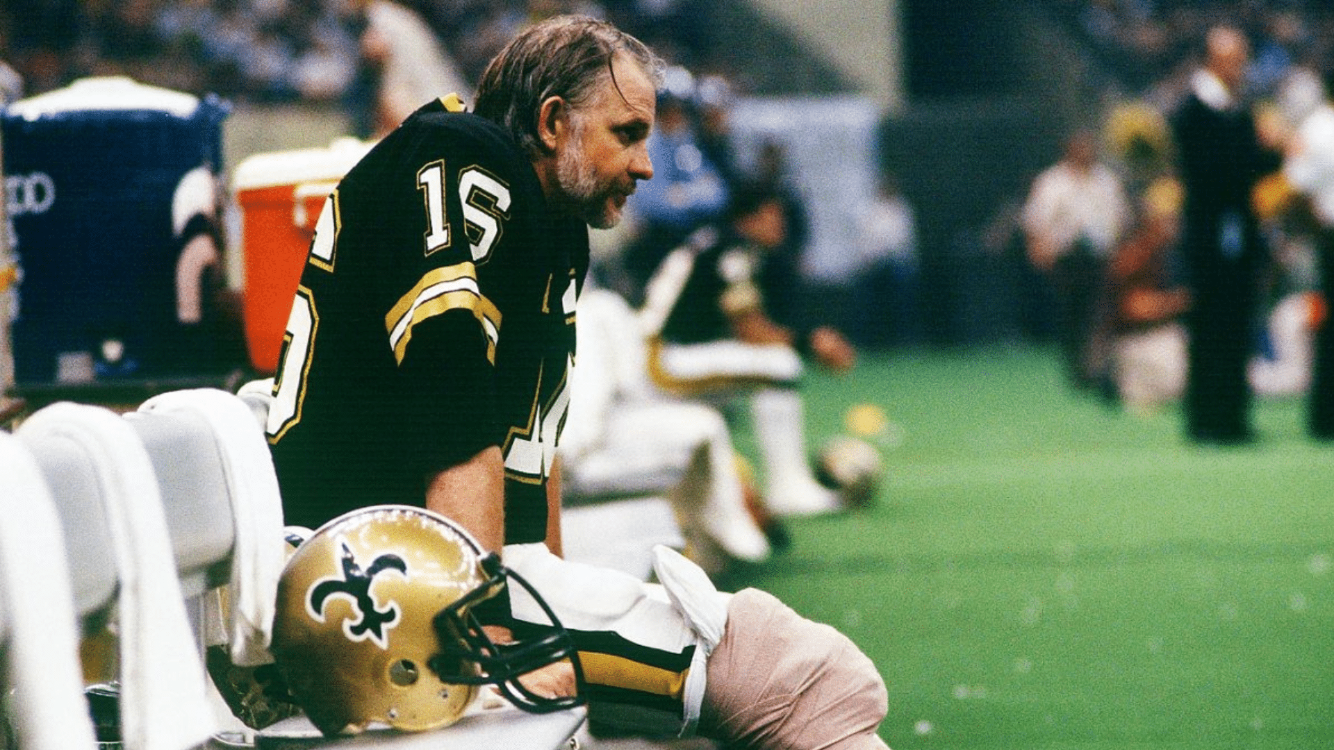 ken stabler best qb of all time sitting on a sideline bench in black and gold football uniform with helmet beside him