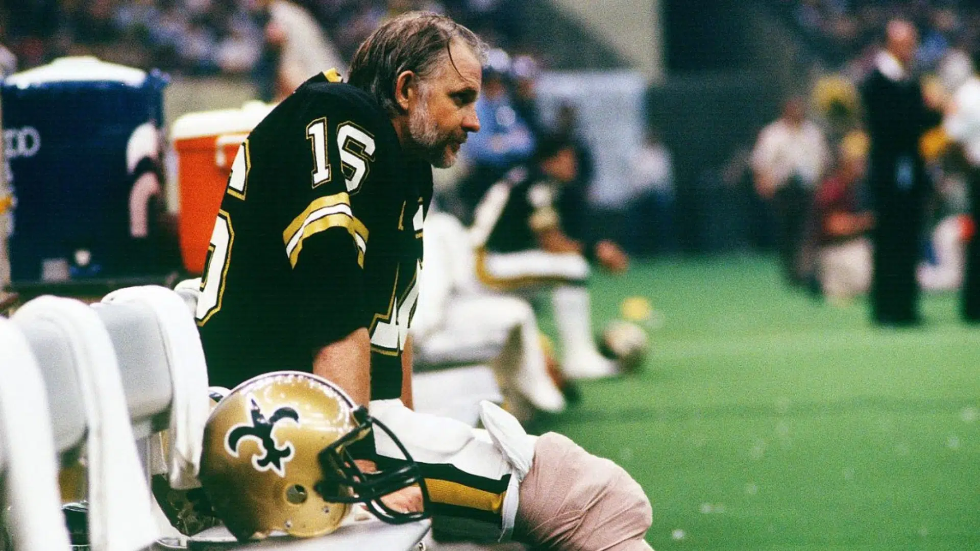 ken stabler best qb of all time sitting on a sideline bench in black and gold football uniform with helmet beside him