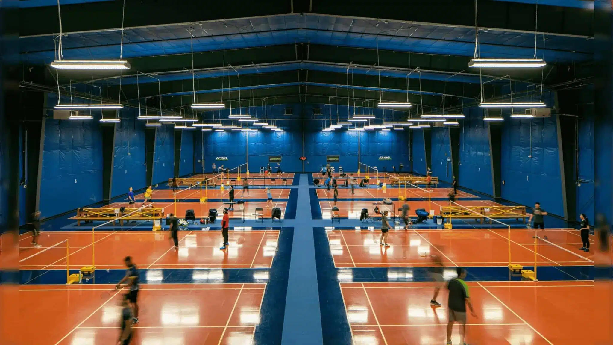 large indoor badminton facility with multiple courts where people are playing matches under bright overhead lights on orange and blue flooring
