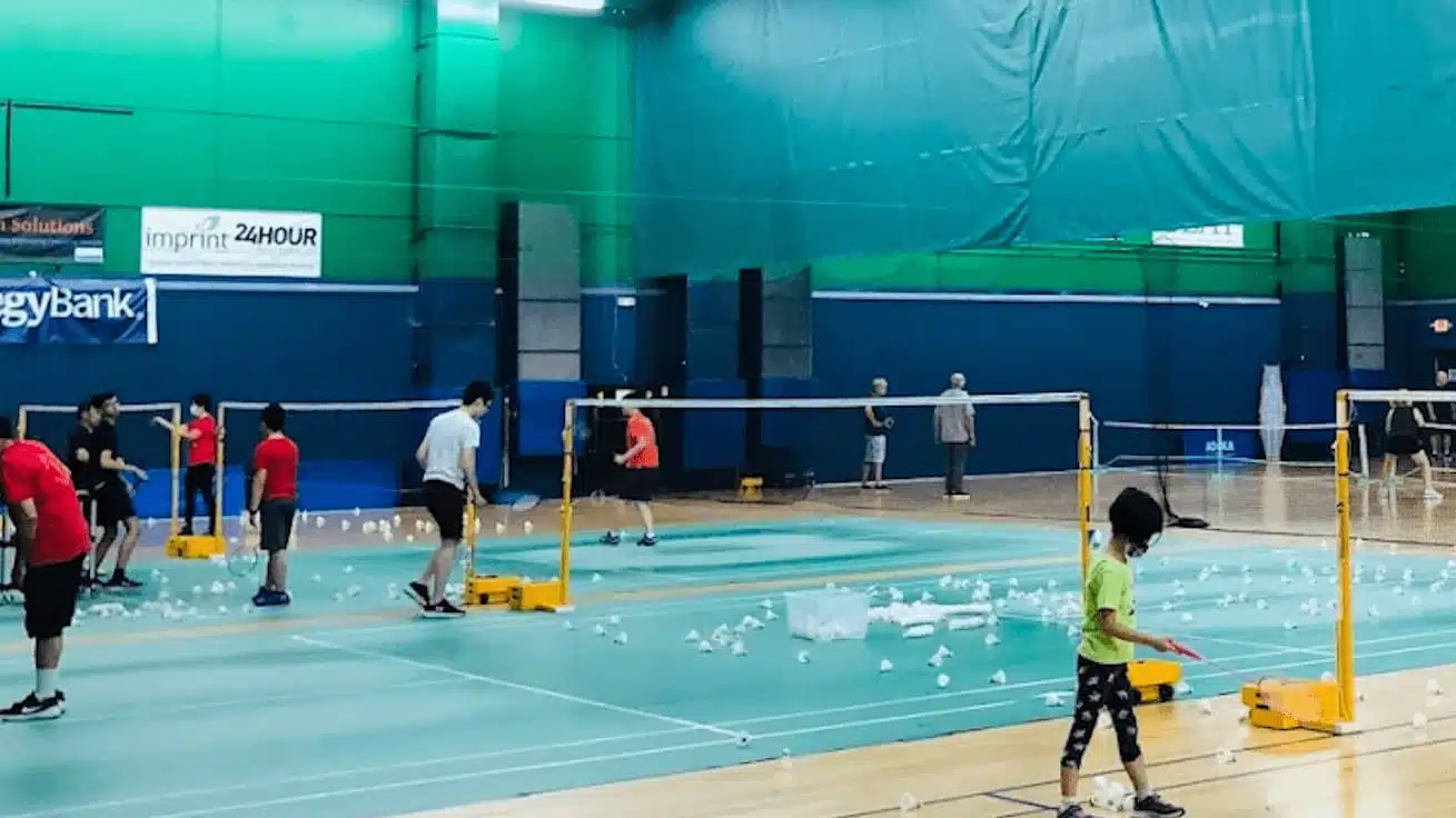 people playing badminton in an indoor gym with multiple courts nets set up and white shuttlecocks scattered across the floor