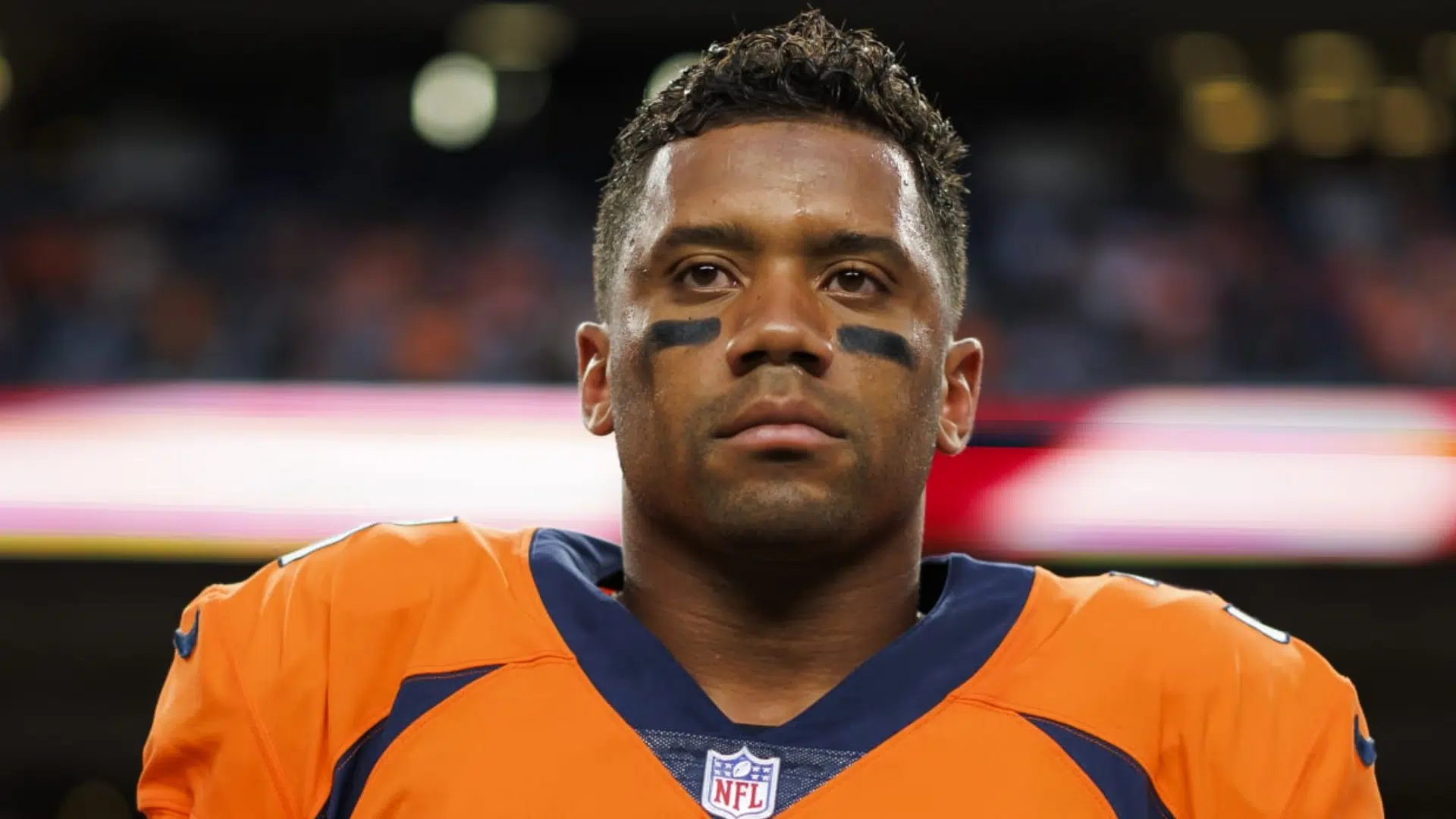 russell wilson focused nfl quarterback stands under stadium lights wearing an orange jersey and eye black
