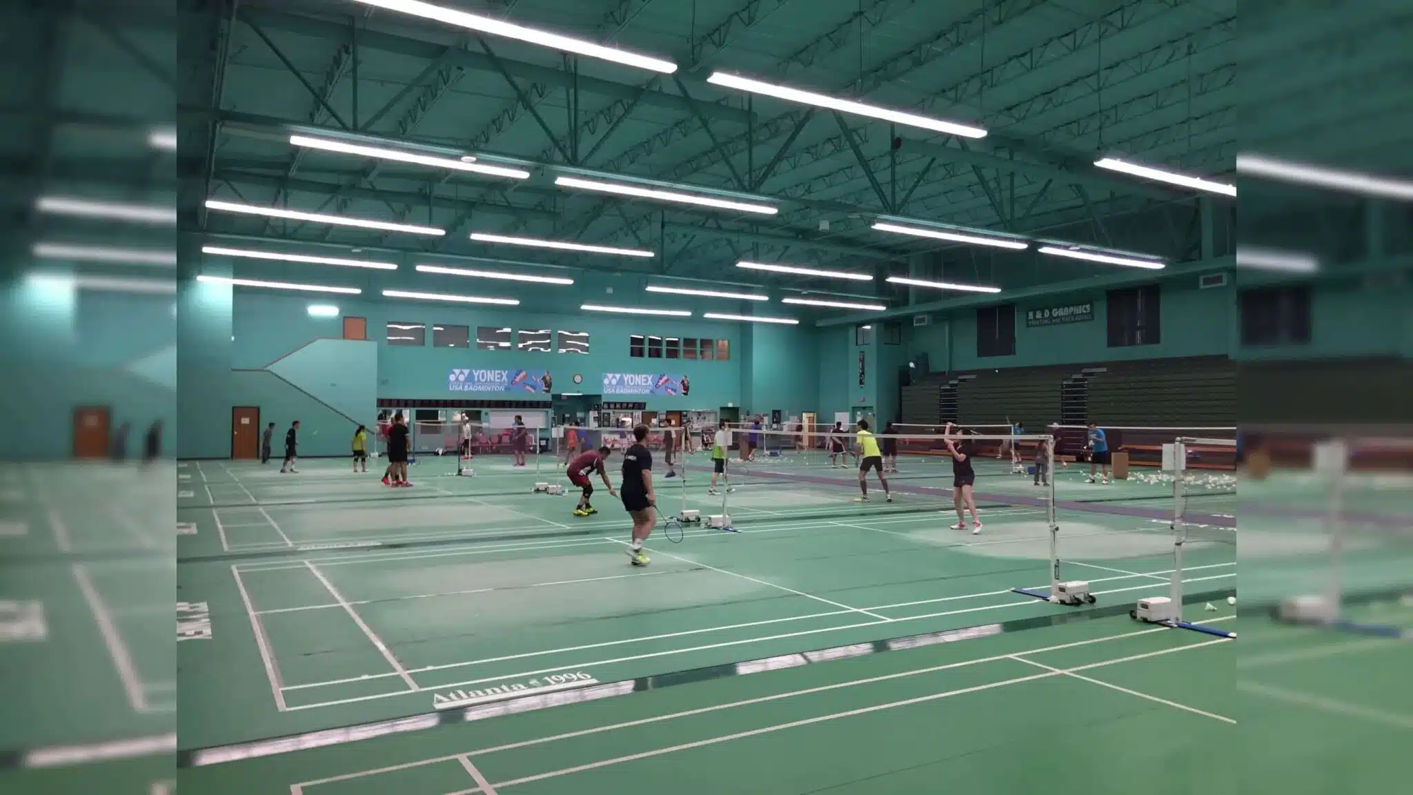 several people playing badminton on multiple indoor courts under bright ceiling lights in a large gymnasium