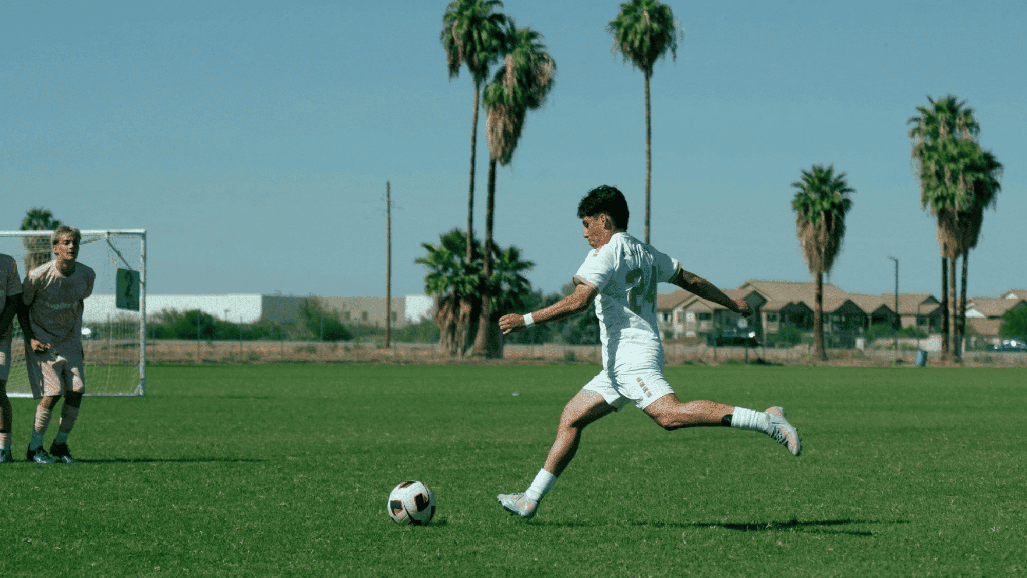 soccer player in white uniform kicks a ball toward goal as goalkeeper watches on a sunny field lined with palm trees