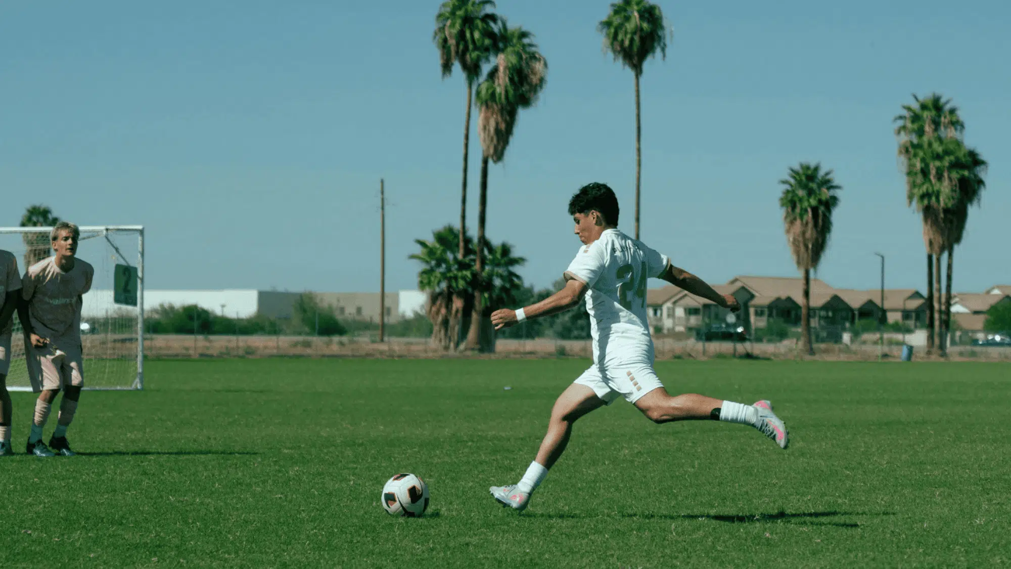 soccer player in white uniform kicks a ball toward goal as goalkeeper watches on a sunny field lined with palm trees