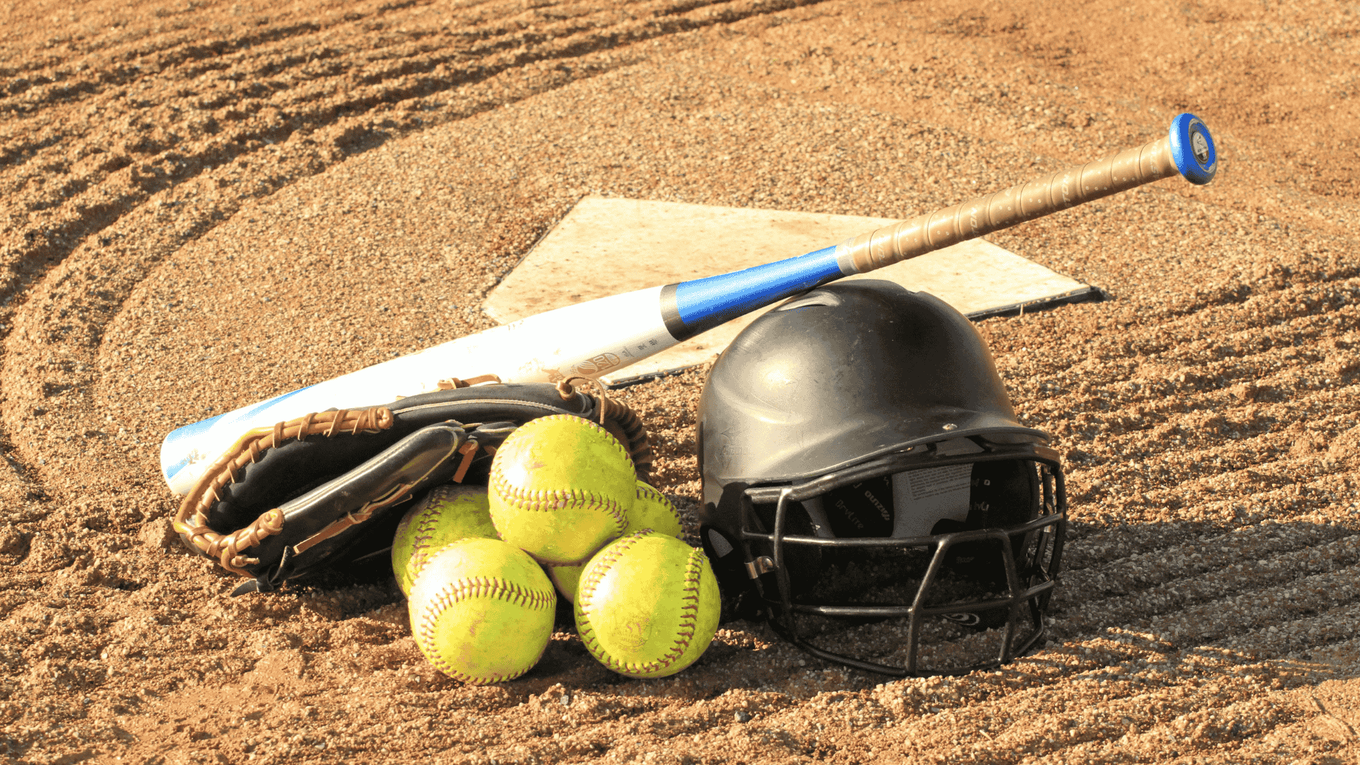 softball bat helmet glove and three balls resting near home plate on a dirt field
