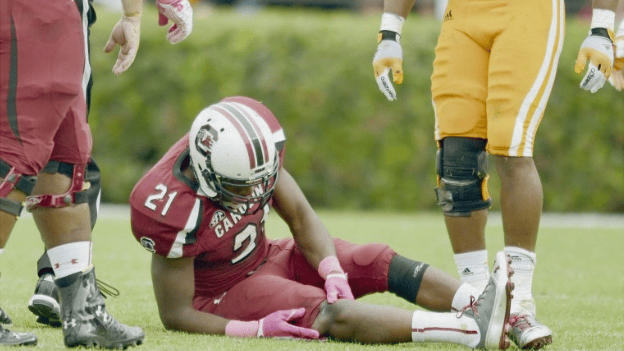 south carolina player marcus lattimore sitting on the field holding his leg while opponents stand nearby